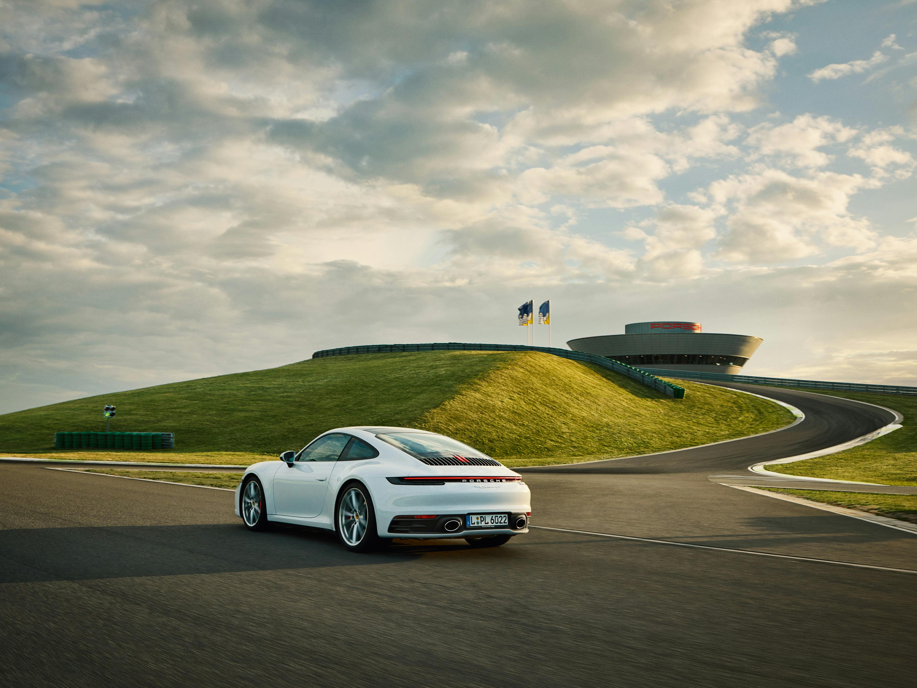 Une Porsche 911 Carrera blanche roule sur la piste d'essai du Porsche Experience Center de Leipzig sous un ciel nuageux spectaculaire.