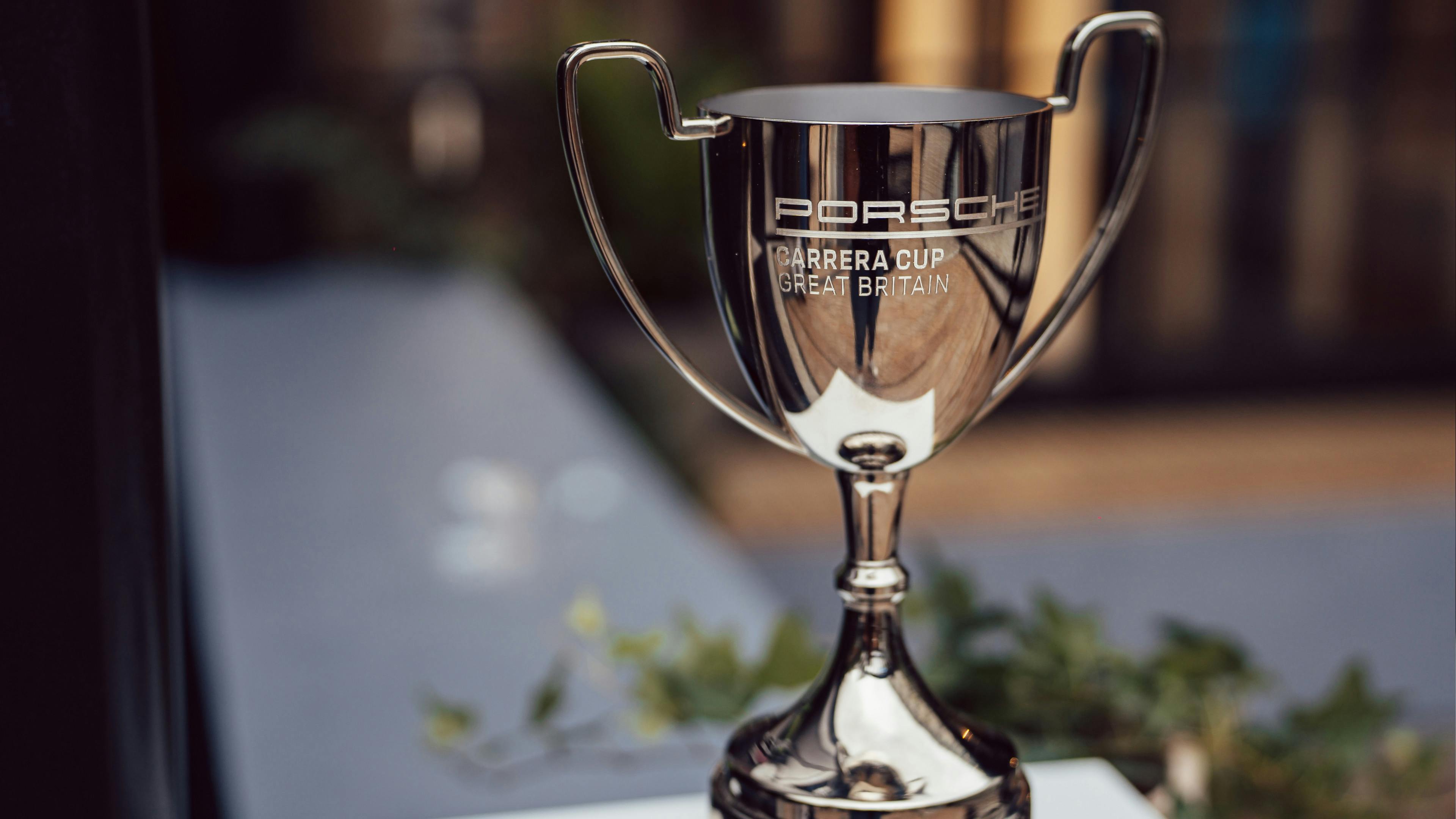 A sharp close-up of a silver trophy engraved with Porsche Carrera Cup Great Britain, standing on a white pedestal with a blurred outdoor background.
