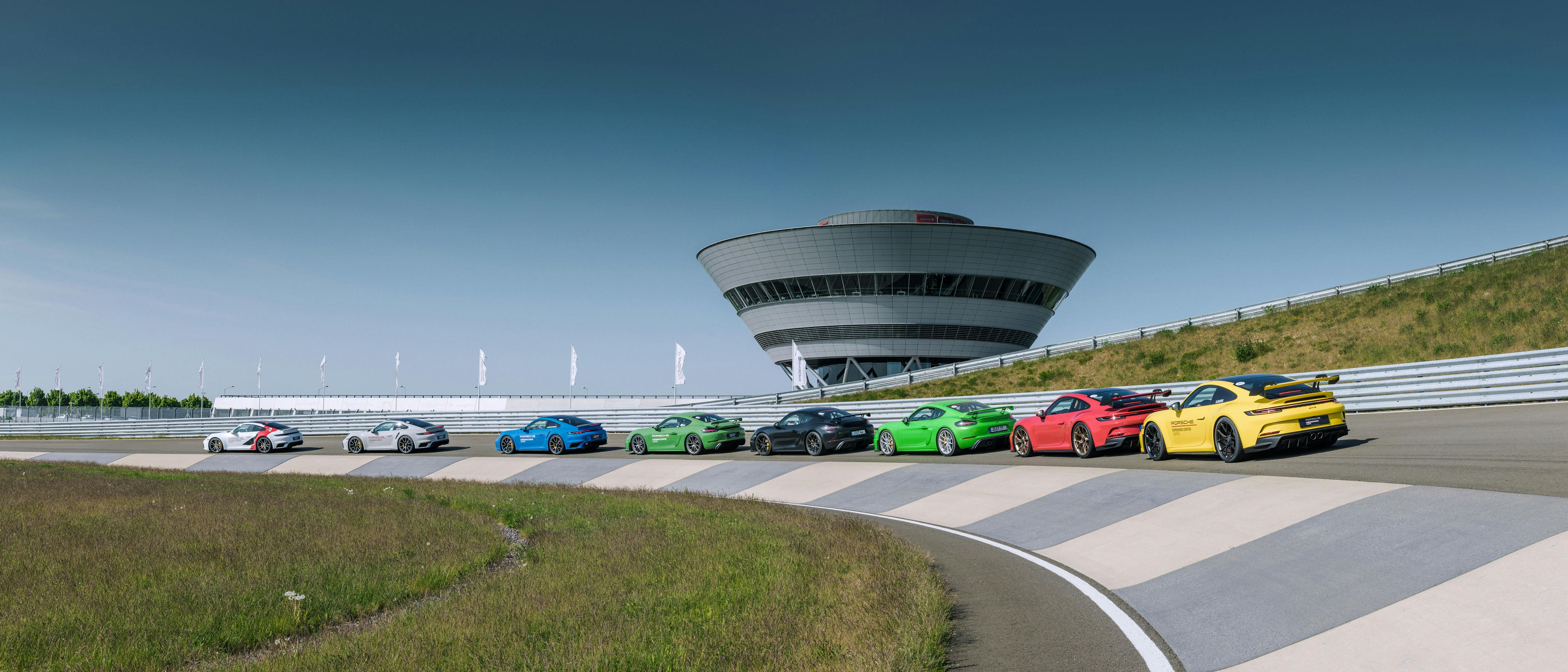 Фотография ряда моделей спортивных автомобилей Porsche разных цветов на кольцевом треке Porsche Experience Center в Лейпциге на фоне башни Porsche.