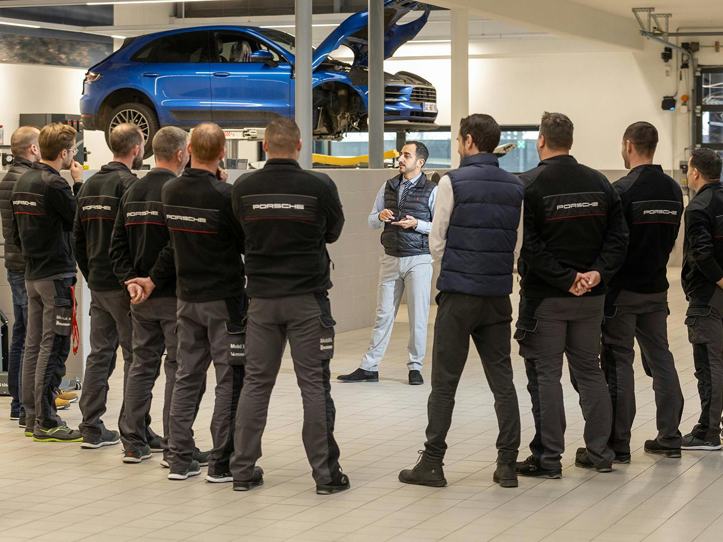 Un groupe de collaborateurs masculins de Porsche se tient en V dans un centre technique Porsche, dos à la caméra, tandis qu'un autre collaborateur de Porsche, situé en face d'eux, explique quelque chose.