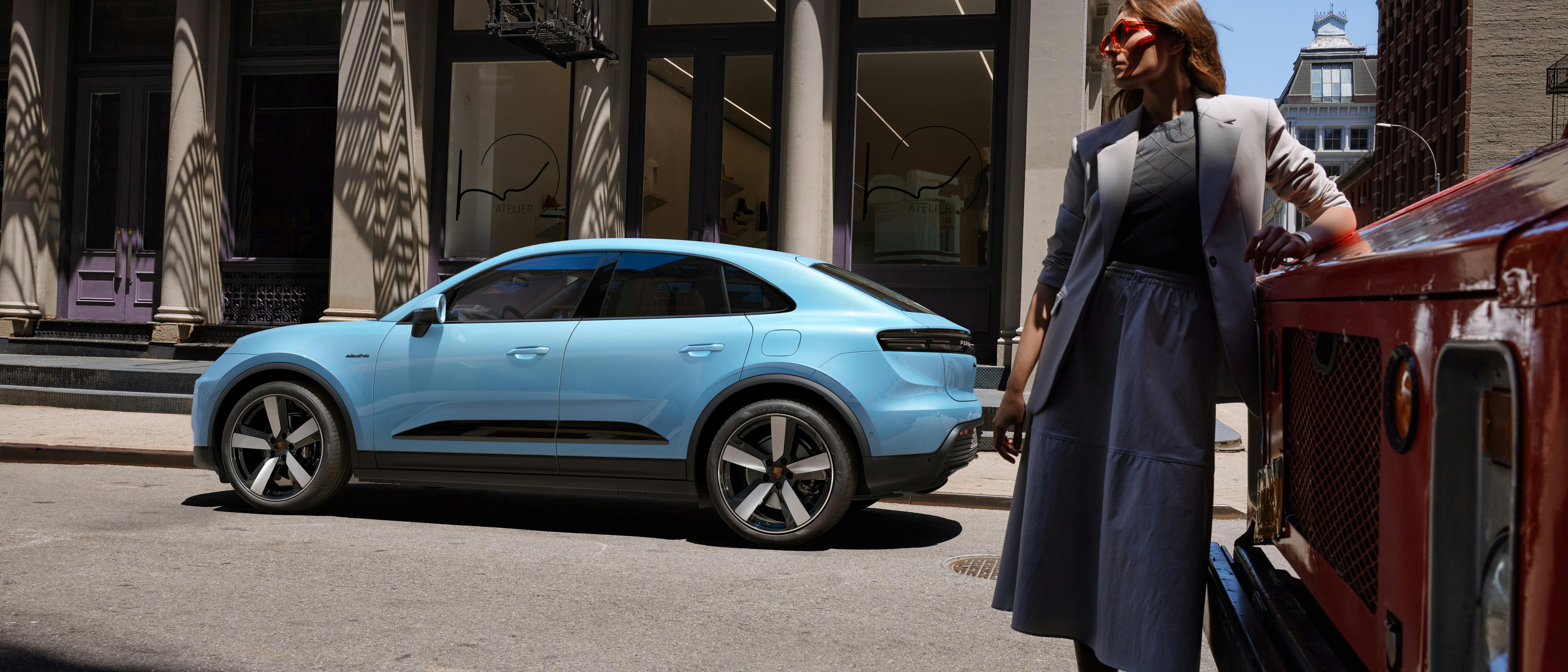 The Macan Electric parked on the side of a road, with a woman leaning on a van in the foreground.
