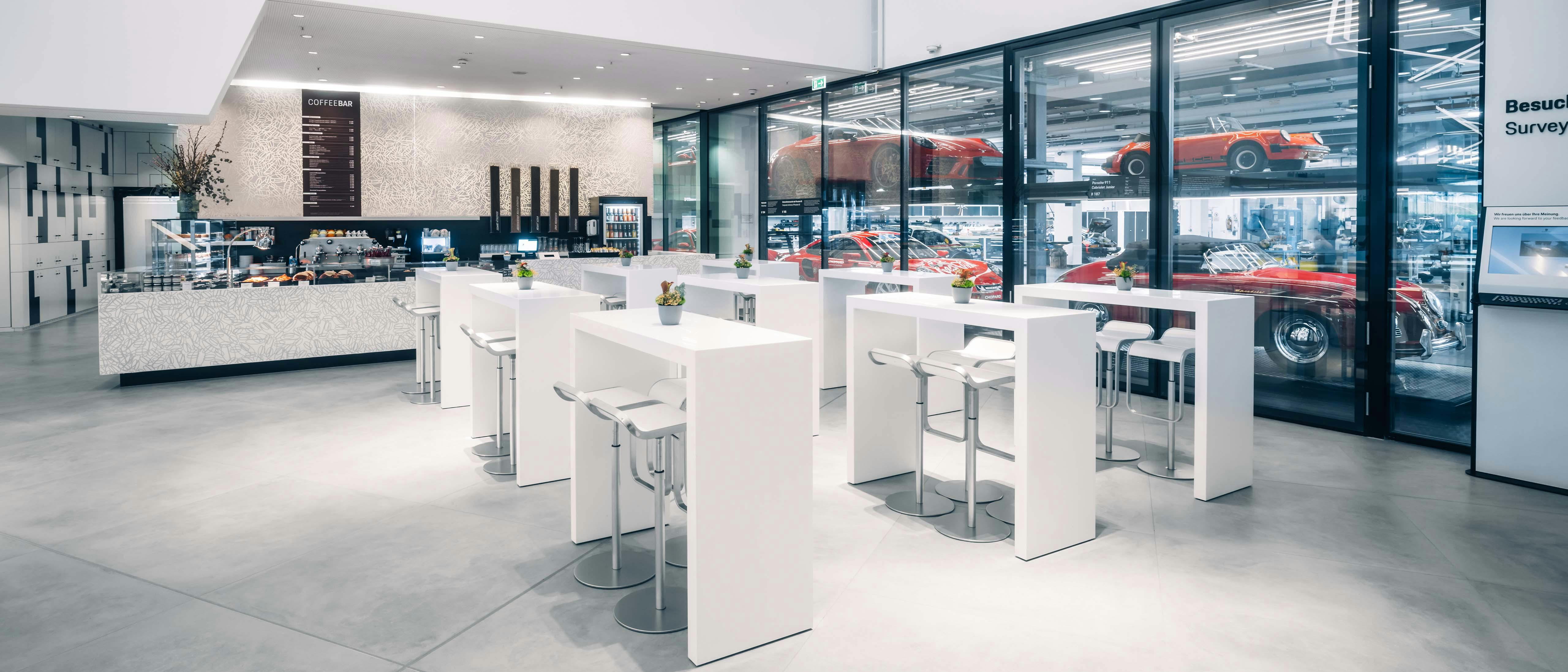 Heller Foyerbereich mit weißer Kaffeebar, hohen Tischen und Blick durch eine Glaswand auf rot lackierte Fahrzeuge in der Werkstatt.