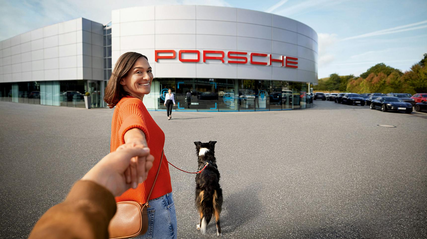 A person in an orange sweater holds hands with someone while walking a dog in front of a Porsche dealership.