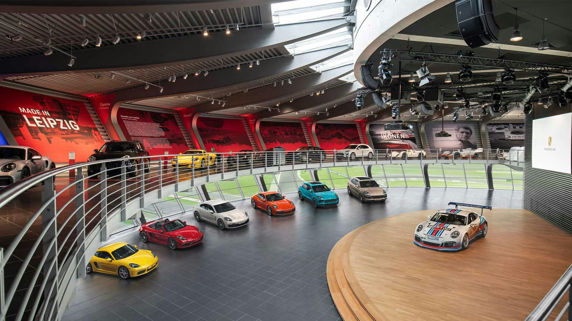 Porsche models are presented in an indoor showroom with a stage and gallery at the Porsche Experience Centre Leipzig.