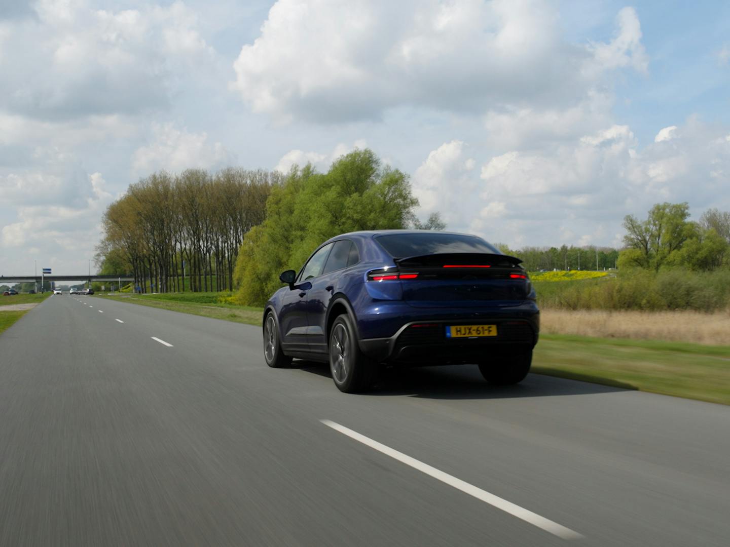 Achteraanzicht van een rijdende donkerblauwe Porsche Macan Electric op een openbare weg door een groen Nederlands polderlandschap met een lichtbewolkte lucht. De foto toont de sportieve lijnen, de actieve achterspoiler en de doorlopende led-achterlichten.