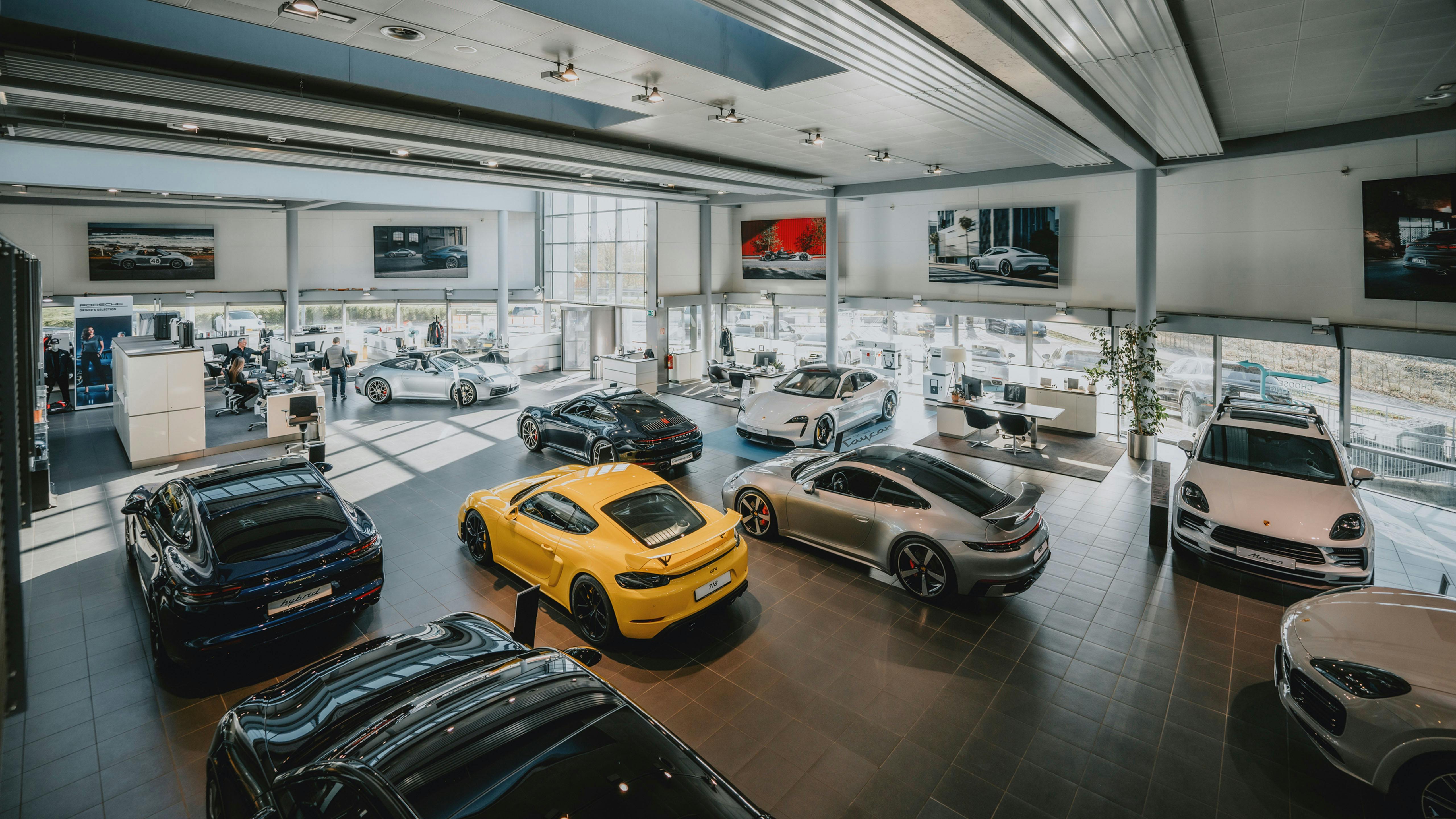 Vue de dessus du showroom du Porsche Zenter Lëtzebuerg avec plusieurs voitures Porsche au sol et des affichages avec des images de Porsche sur les murs au-dessus de la façade vitrée continue.