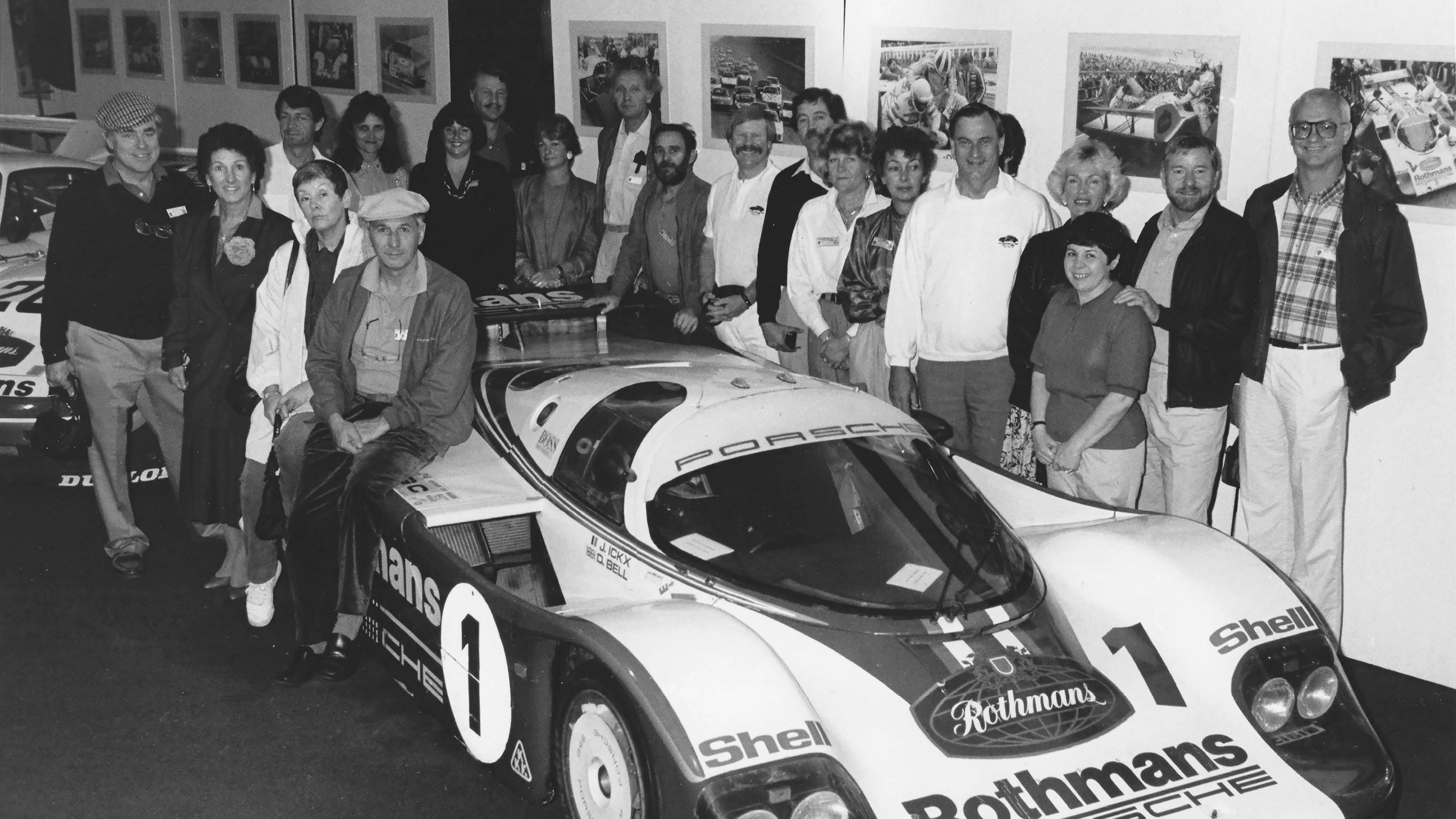 Historic black-and-white photo of a group gathered around a Porsche race car, with motorsport images displayed on the walls.