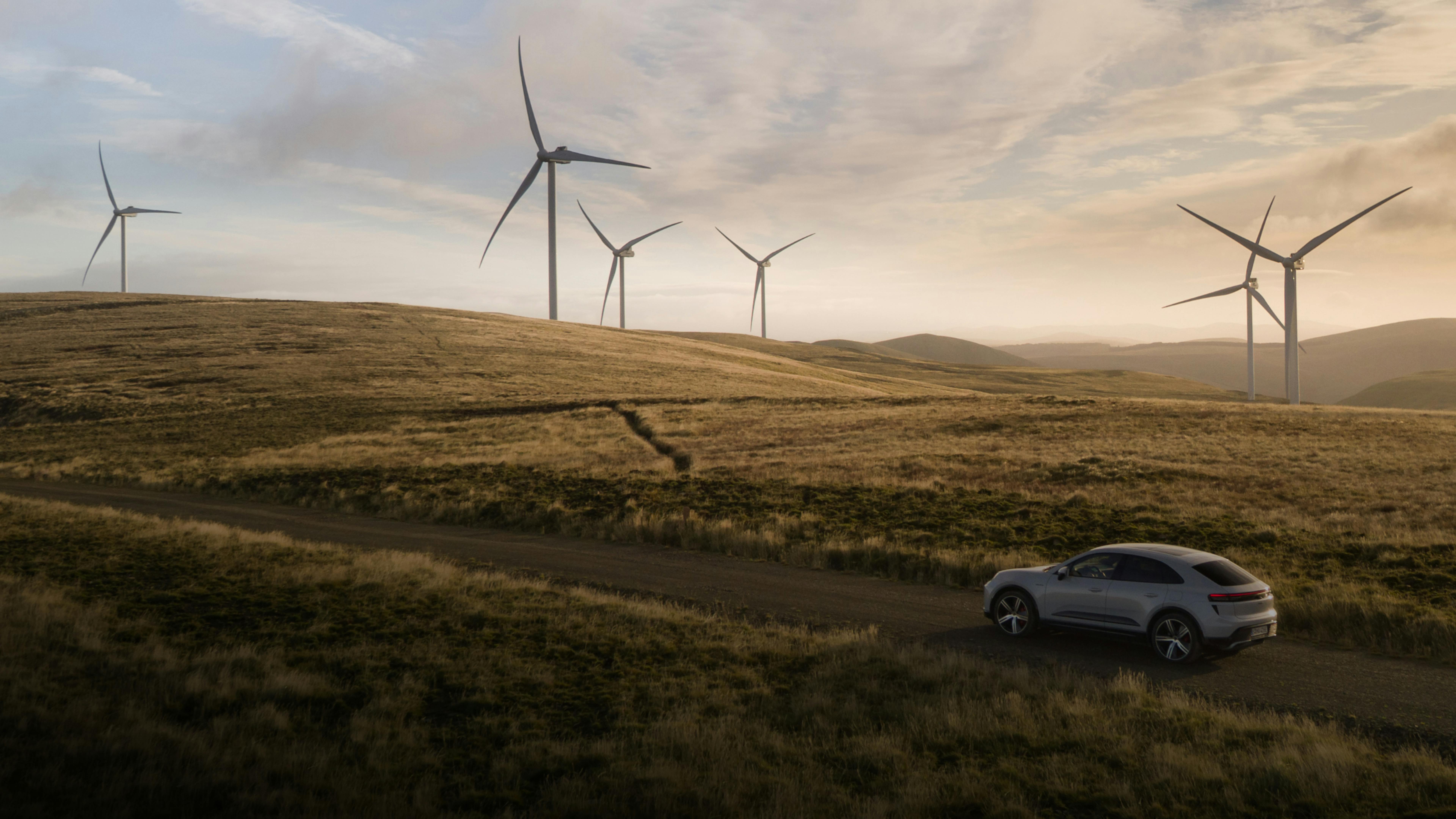 A Porsche Macan Turbo in Ice Grey Metallic driving along a gravel road through a hilly grassy landscape. There are wind turbines in the background.