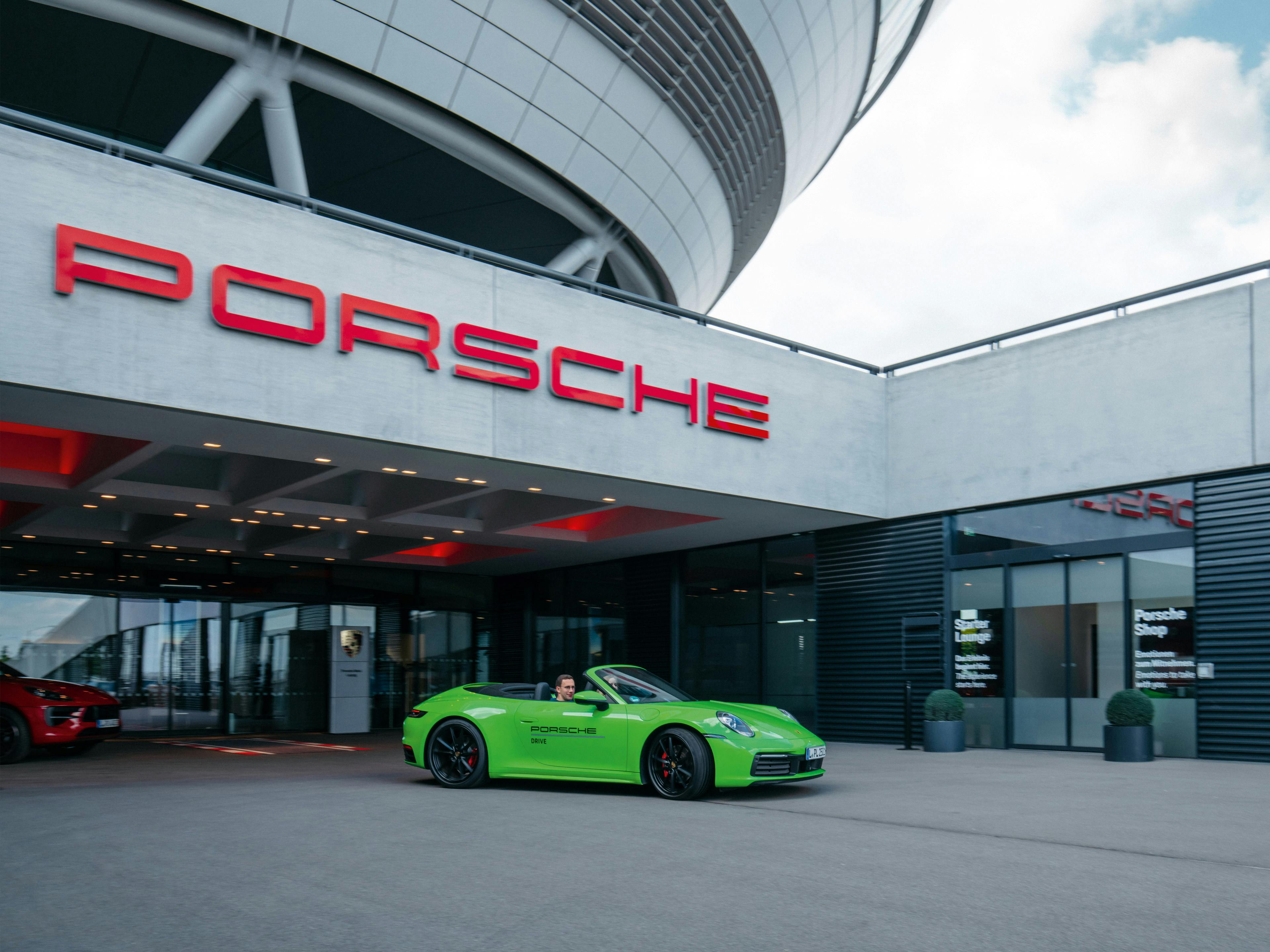 Ein grüner Porsche mit dem Seitenschriftzug Porsche Drive steht vor einem Porsche-Gebäude.