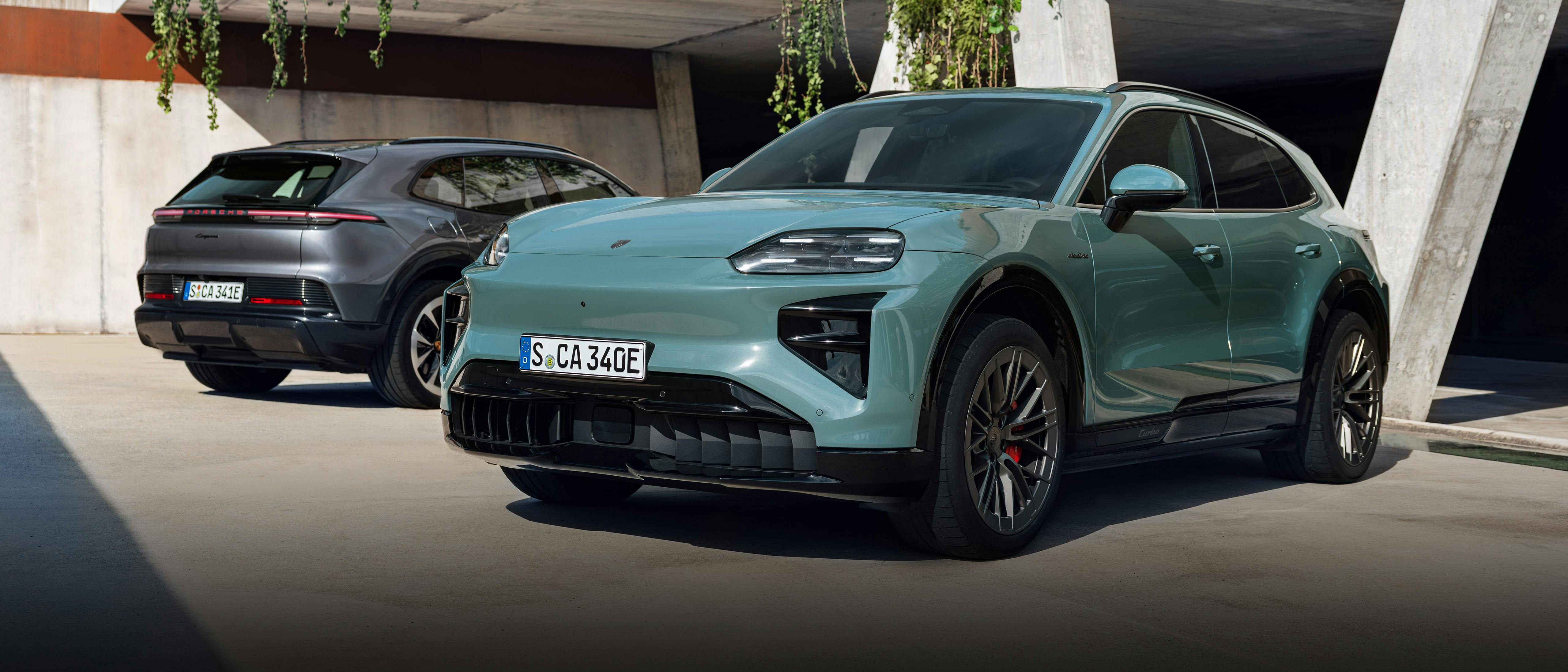 Two Porsche electric SUVs parked outside a modern concrete building with hanging greenery; a light green Porsche Cayenne Turbo Electric is in the foreground, and a dark grey Porsche Cayenne Electric is in the background.