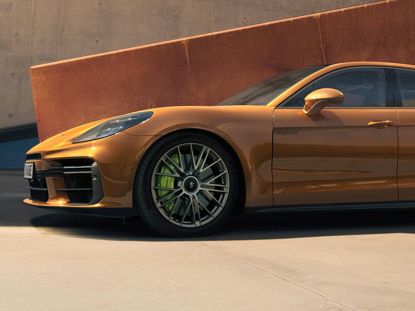 A Panamera Turbo E-Hybrid in Madeira Gold Metallic parked alongside a wall that is copper coloured while there is a lighter brown building behind it.