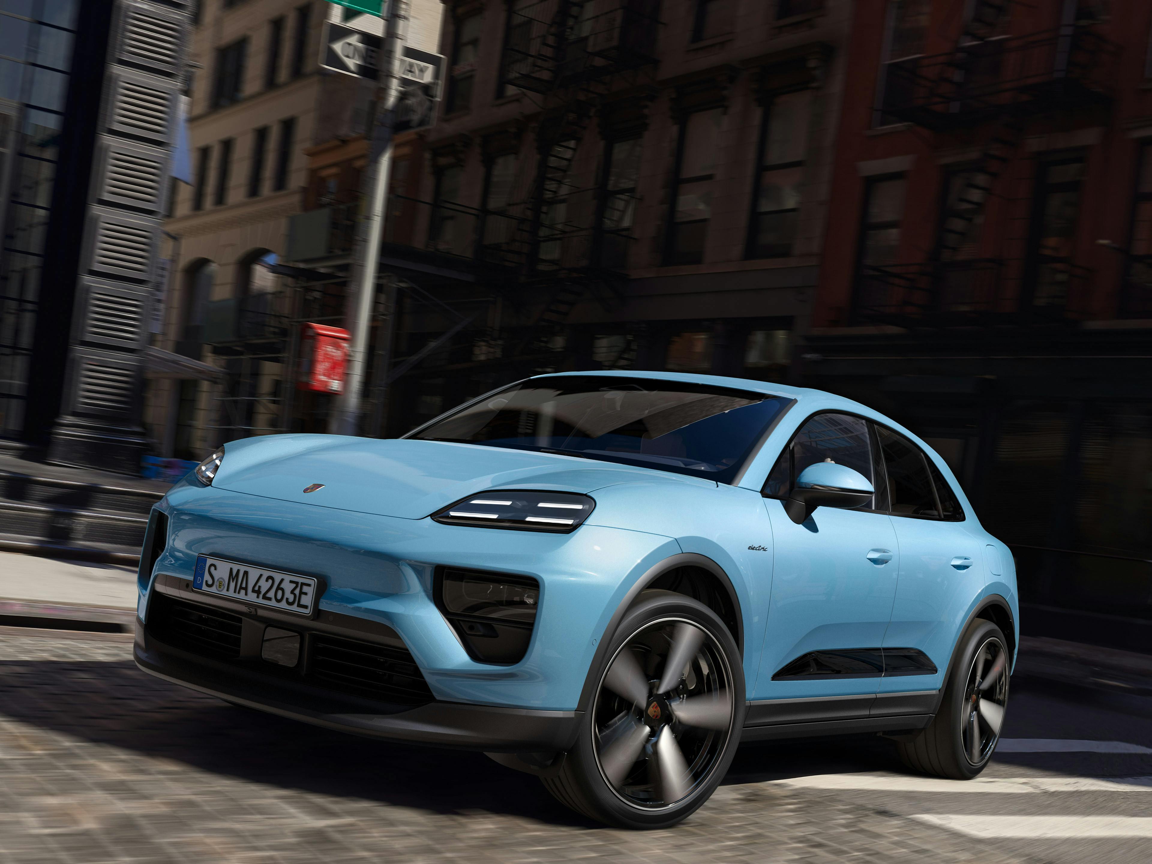 Zwei Porsche Macan Elektro, einer weiß und der andere lila, sind unter einer eisernen Brücke geparkt, mit einer Stadtansicht im Hintergrund.