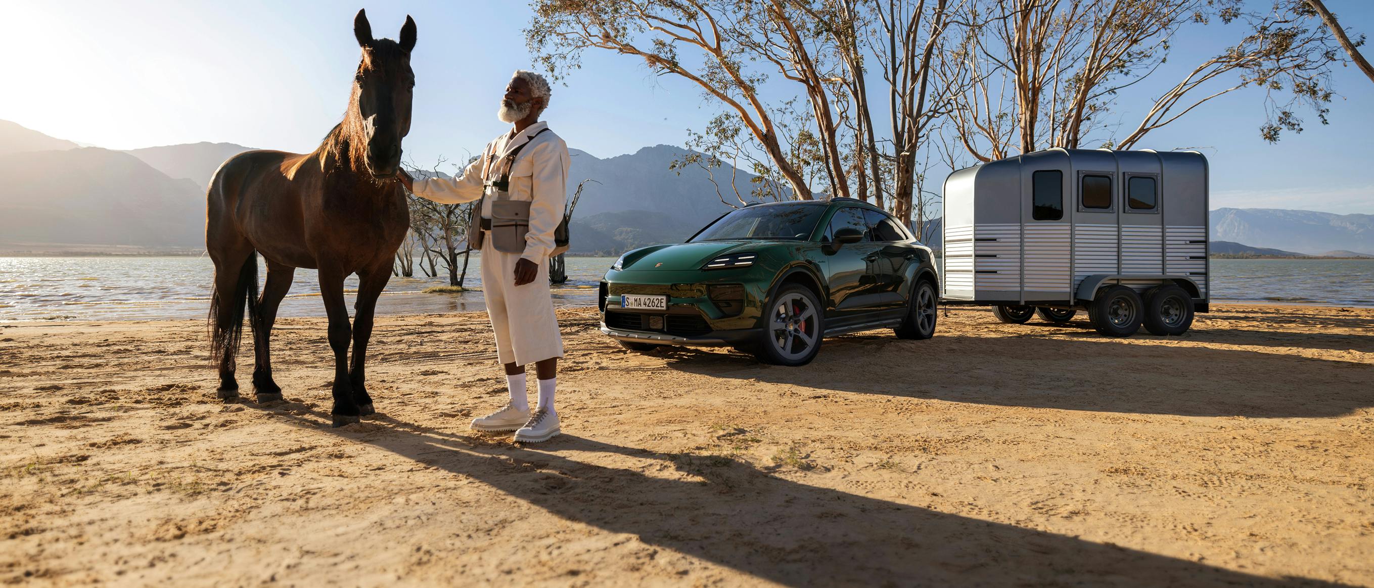 Um homem junto a um cavalo num ambiente desértico. Em segundo plano, um Porsche Macan 4S com um reboque para cavalos.