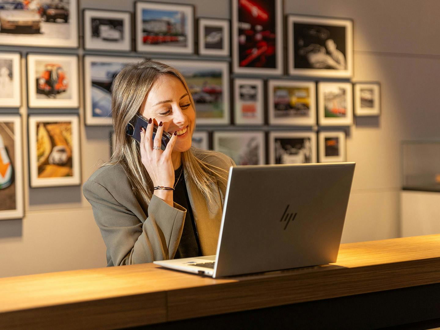 Une employée de Porsche, assise derrière un comptoir en bois, écoute quelqu'un sur son smartphone tout en regardant l'ordinateur portable ouvert sur le comptoir.