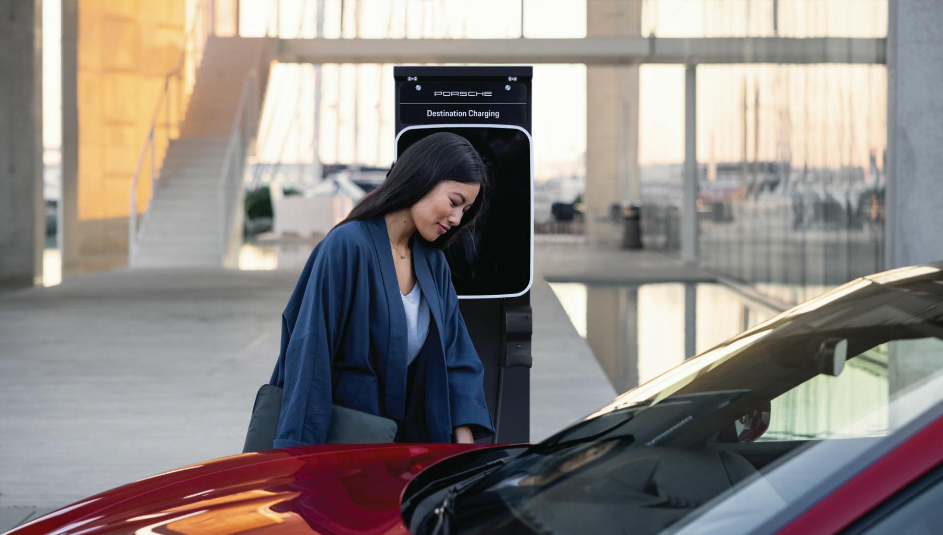 A woman stands at a public charging pedestal and charges her Porsche Taycan.