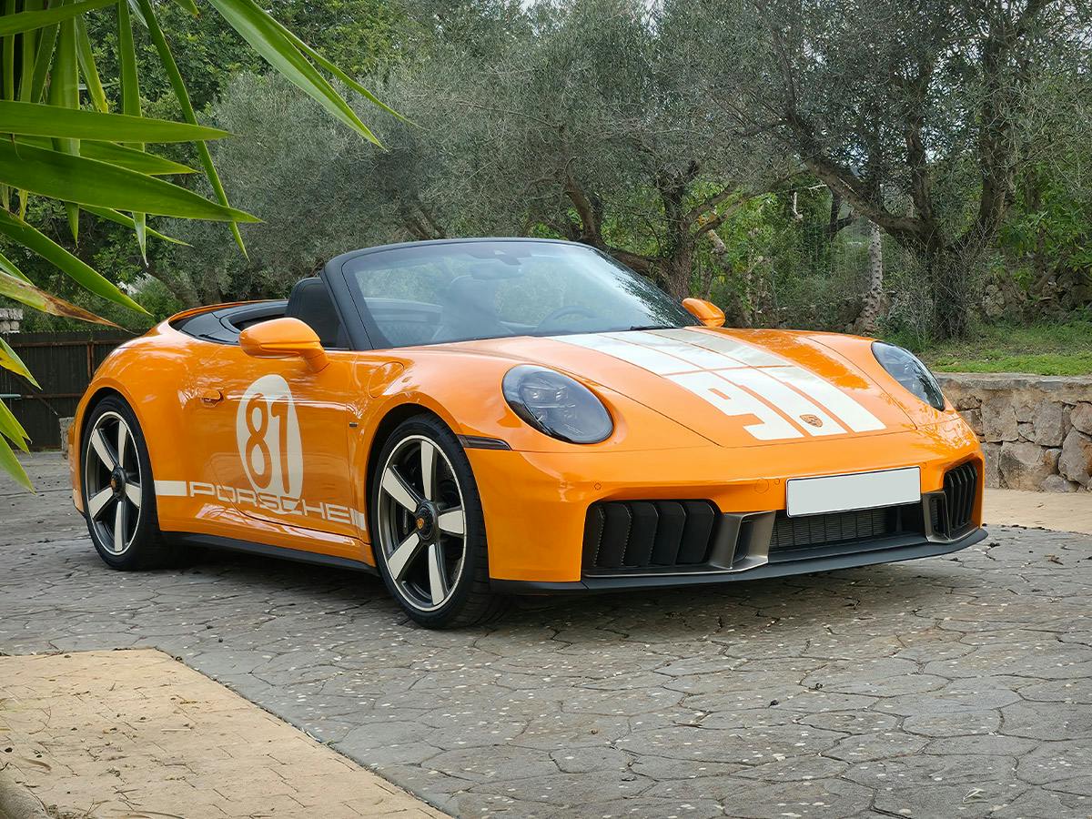 Porsche 911 Spirit 70 in Paint to Sample colour Signalorange, shown from a front-side angle, featuring individualised wheels and decals on the side and bonnet, parked on a stone-paved driveway with trees and greenery in the background.