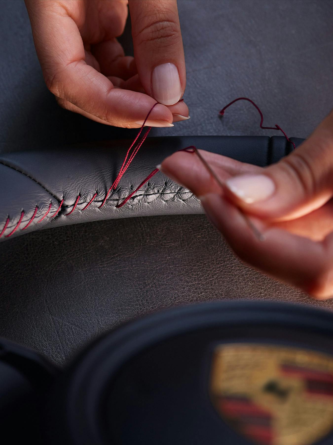 Close-up of a craftsperson hand-stitching red thread into the leather steering wheel of a 911, with the Porsche crest visible below.​
