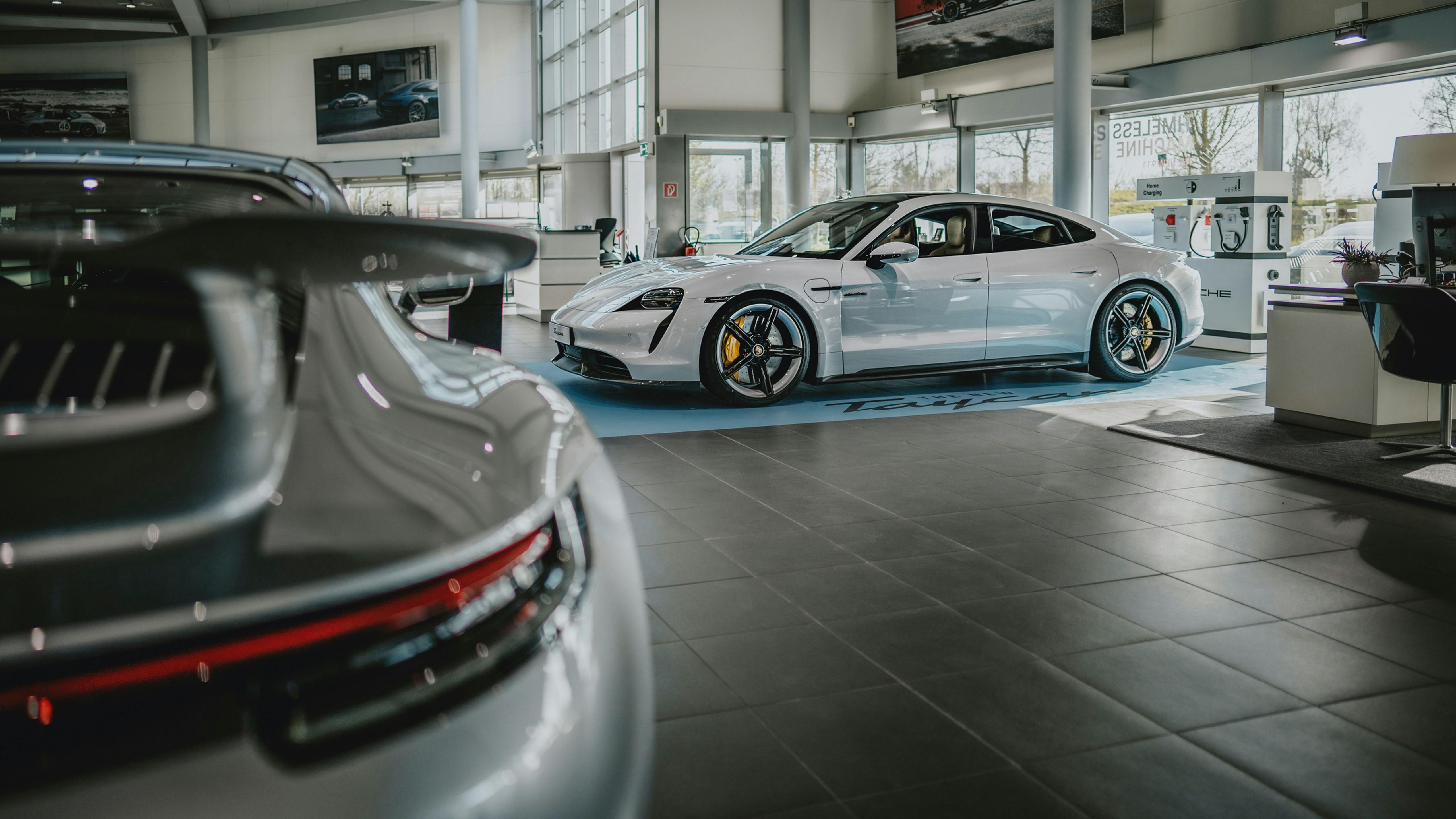 Vue latérale d'un modèle Taycan dans le showroom du Porsche Zenter Lëtzebuerg, mis en valeur par un carré bleu clair au sol avec l'inscription "La nouvelle Taycan" et une borne de recharge électrique factice derrière la voiture.