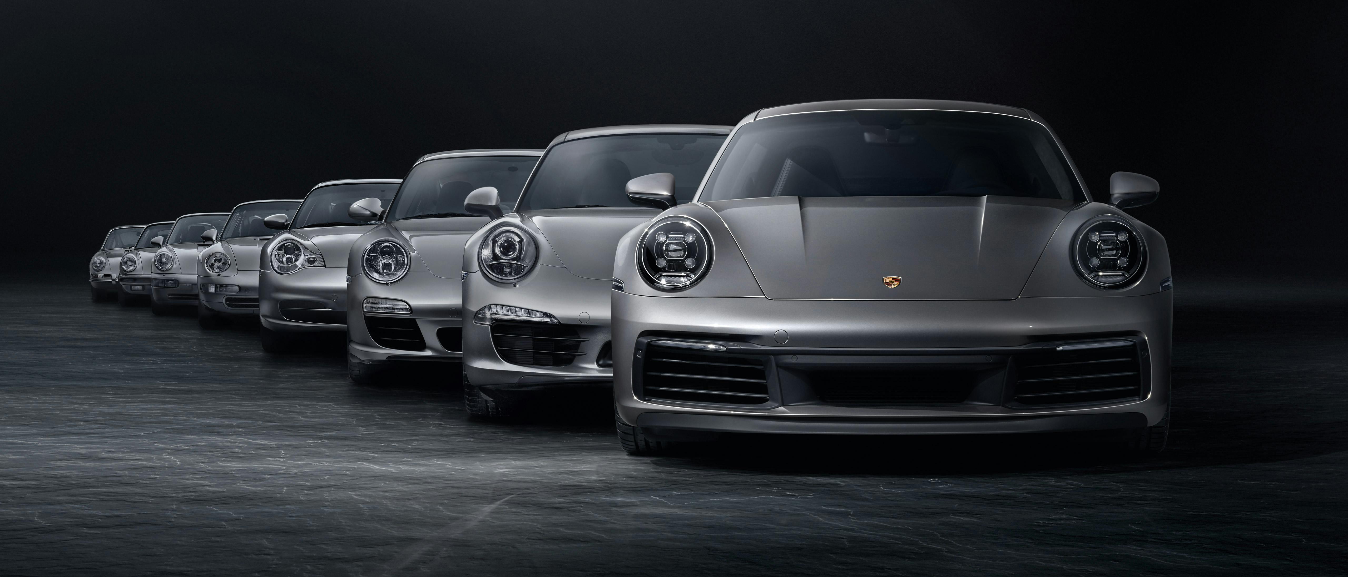 In einem schwarzen Studio stehen verschiedene silberne Porsche 911 Generationsmodelle hintereinander in einer Reihe.