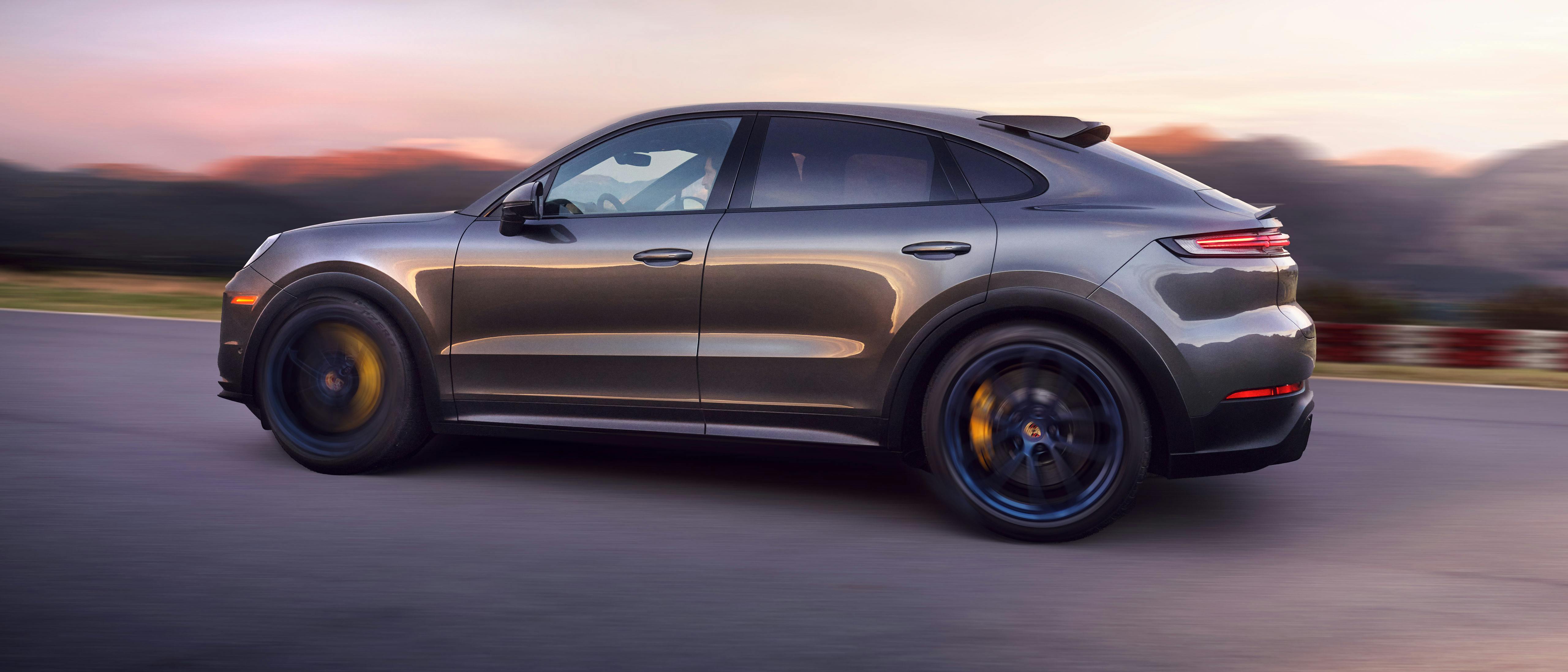 A sleek, dark gray Porsche SUV driving on a road at sunset, showcasing its sporty and aerodynamic design.
