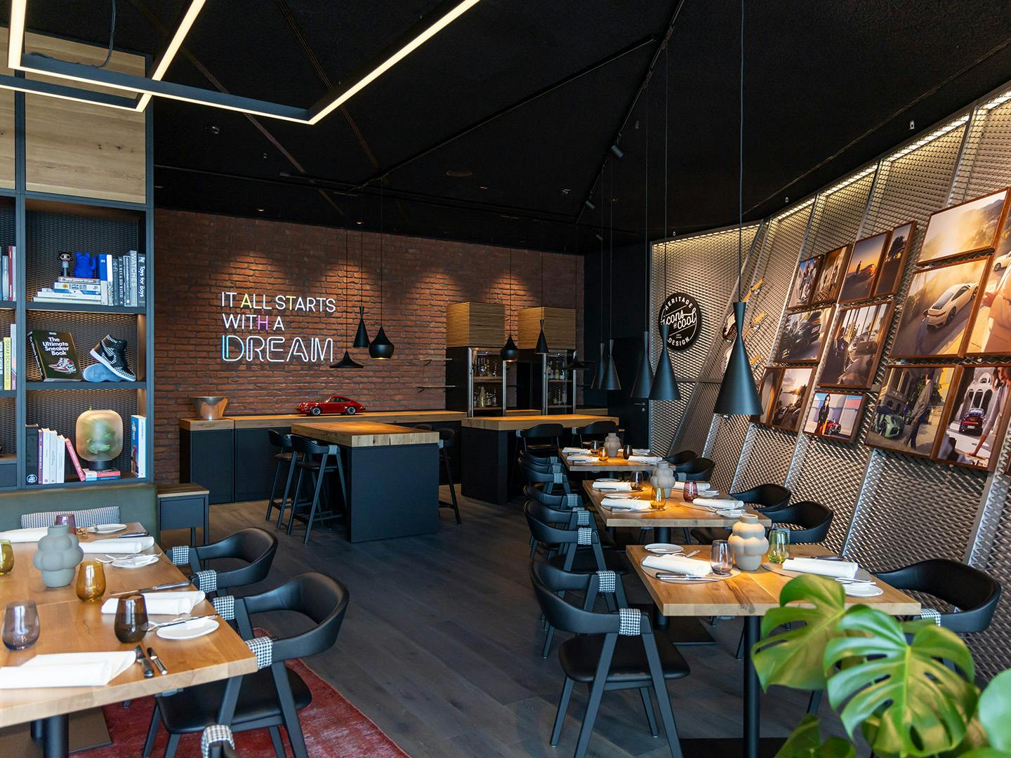 Loungebereich im Porsche Museum, angrenzend an das Restaurant Christophorus: stilvoll eingerichteter Raum mit gedeckten Tischen, Backsteinwand mit dem Schriftzug ‚IT ALL STARTS WITH A DREAM‘, gerahmten Fotografien an der rechten Wand sowie Bü-cher- und Dekorelementen auf Regalen links – warmes Licht schafft eine einladende Atmosphäre.
