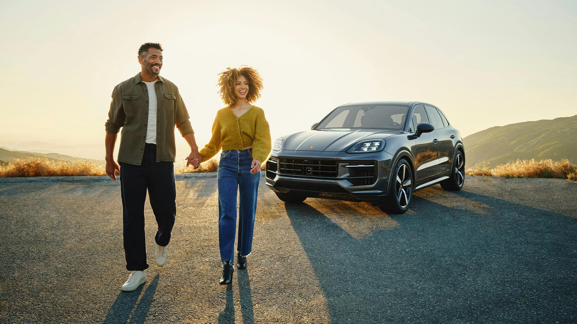 Un couple marche main dans la main devant une Porsche sur une route au coucher du soleil.
