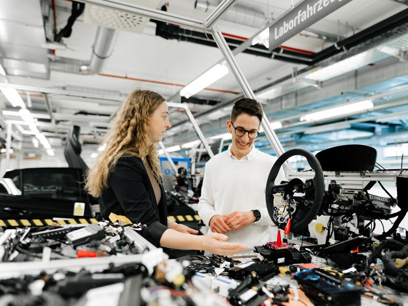 Zwei junge Studierende bei Porsche stehen an einem Laborfahrzeug und sie erläutert Funktionen.