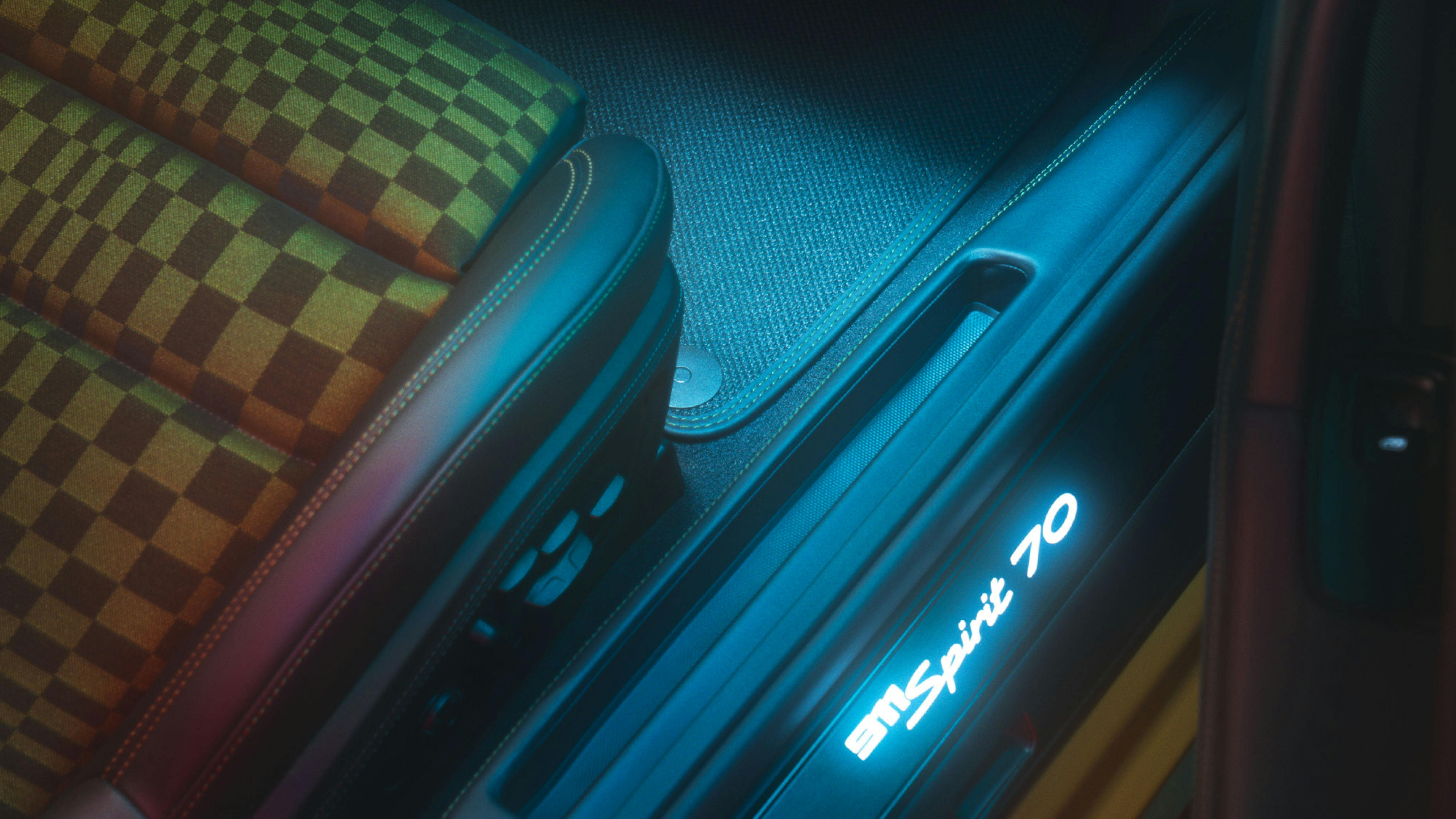 Detalle del interior de un Porsche 911 70 Spirit, mostrando el asiento con diseño a cuadros, la iluminación ambiental azul y el logotipo "911 Spirit 70" iluminado en el umbral de la puerta.