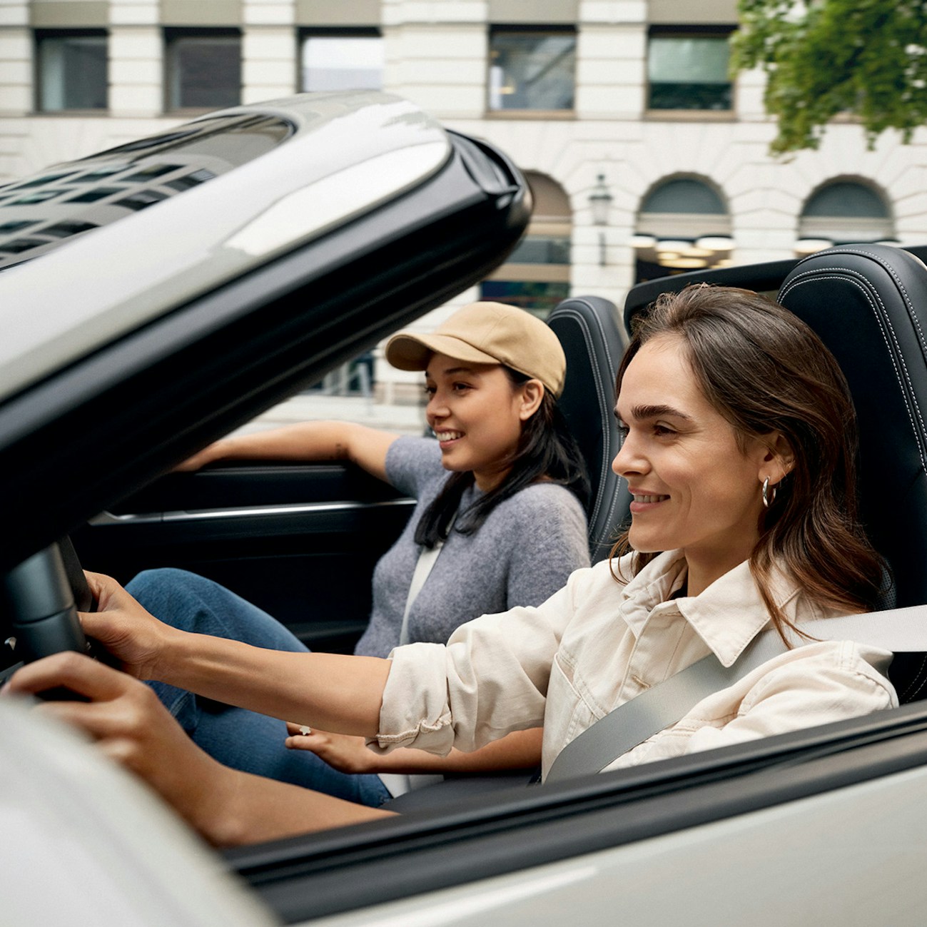 Two smiling young women driving a Porsche car on an urban street.