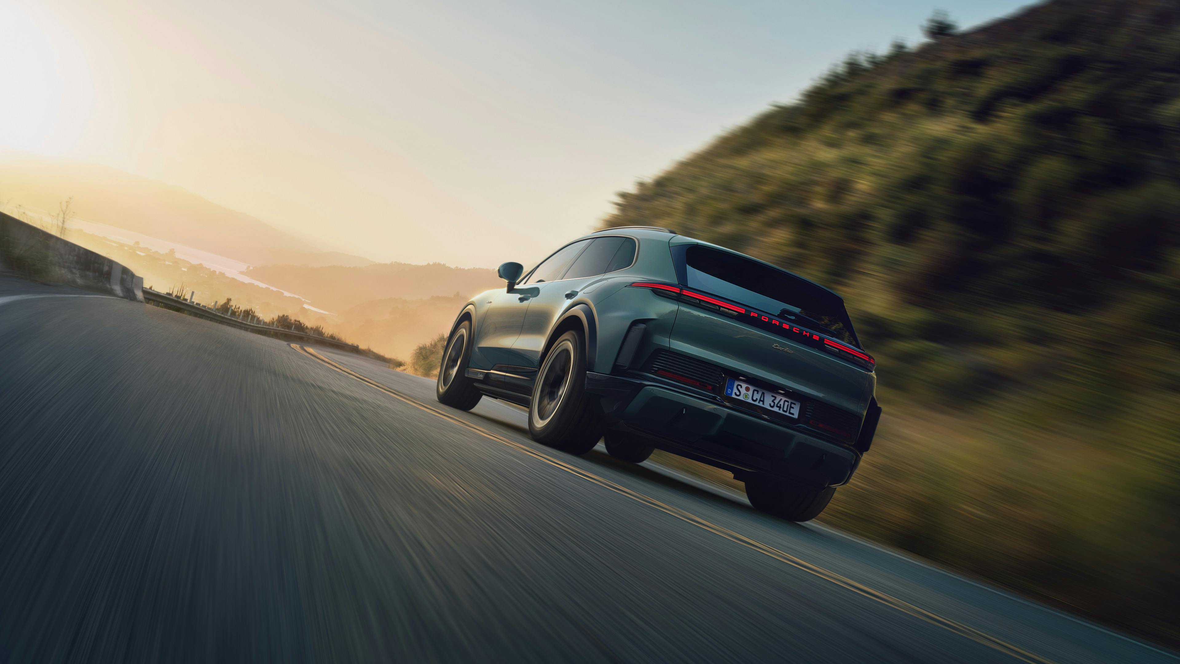 A Mystic Green Metallic Porsche Cayenne Electric driving on a winding mountain road. The image is blurred to emphasize motion.