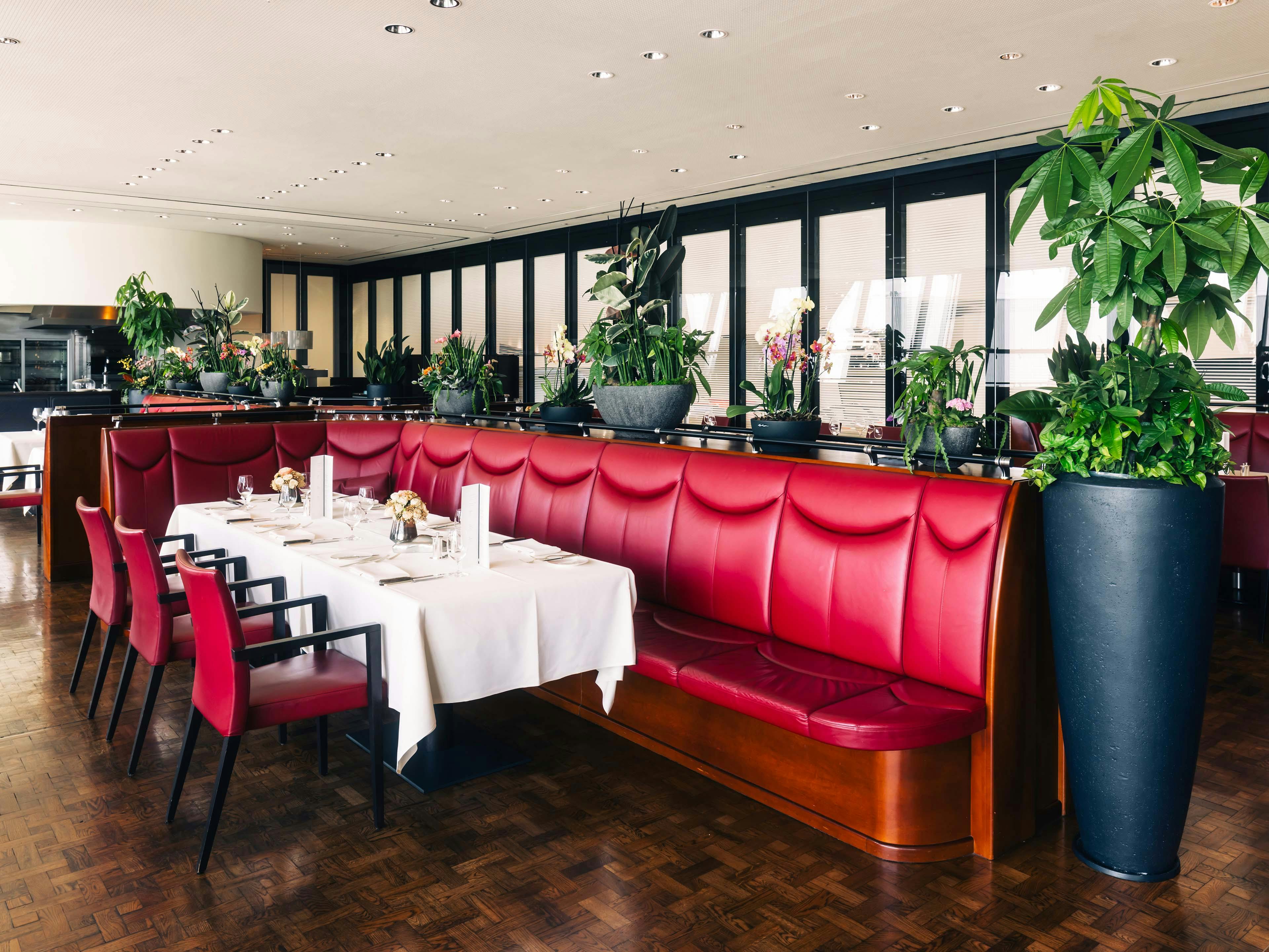 Innenraum des Restaurantbereichs mit roten Polsterbänken, gedeckten Tischen und großen Pflanzen.