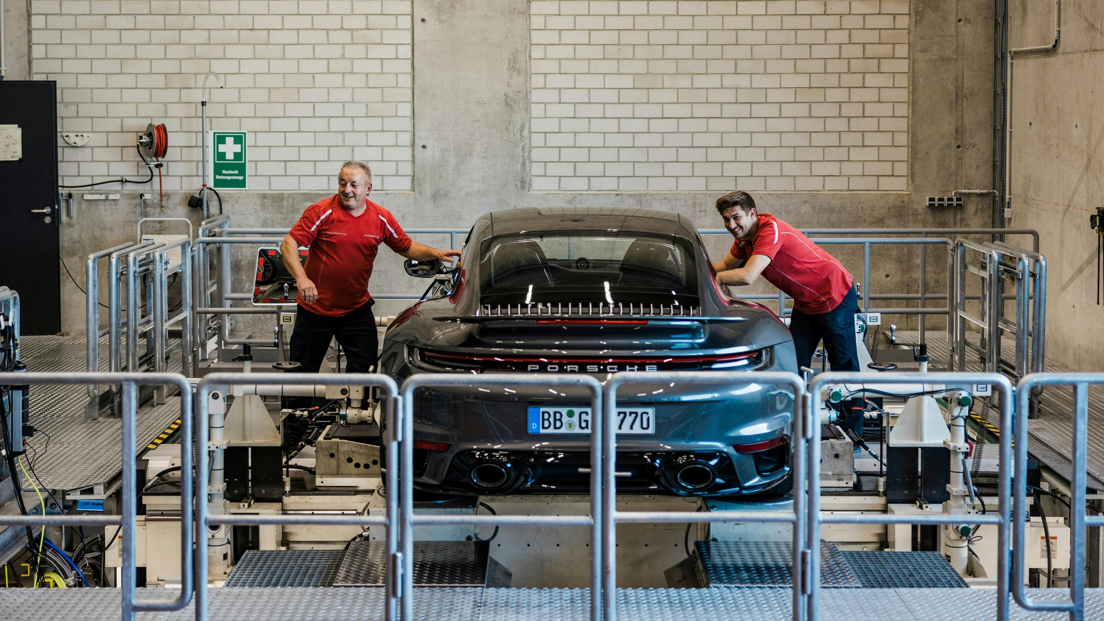 Zwei Personen in roten Porsche‑Arbeitsshirts arbeiten an einem schwarzen Porsche‑Sportwagen auf einem Prüfstand in einer technischen Werkstatt mit Absperrungen, Messgeräten und industrieller Umgebung.