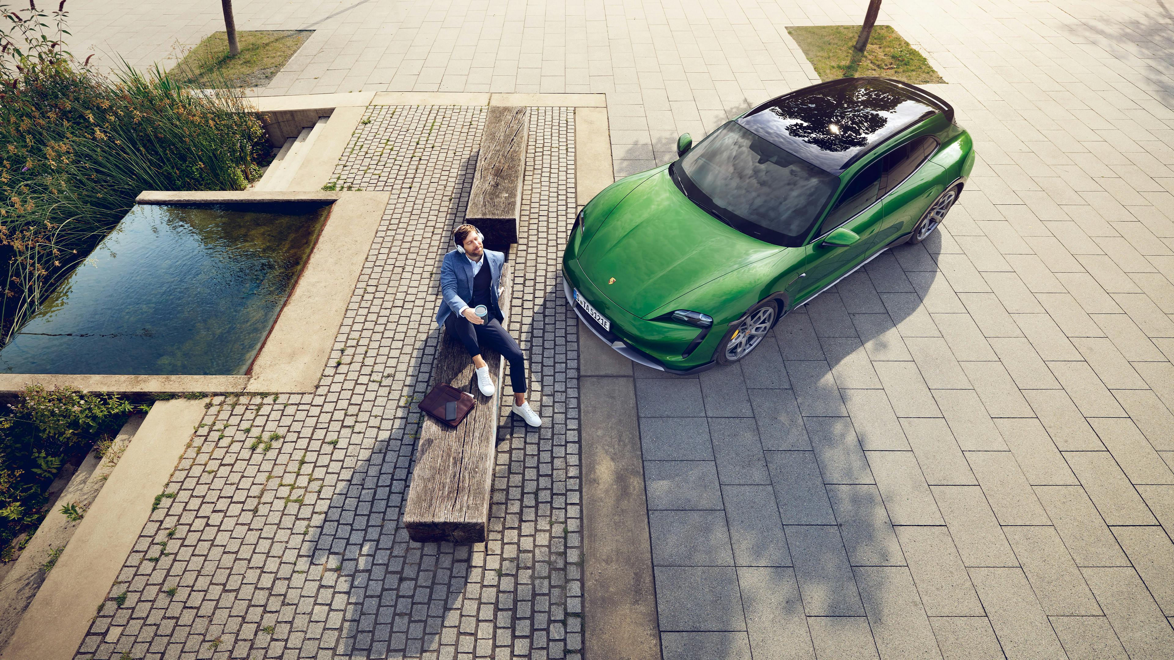 Ein Mann mit Coffee-to-Go-Becher sitzt auf einer Bank neben einem Brunnen, daneben parkt ein grüner Porsche.