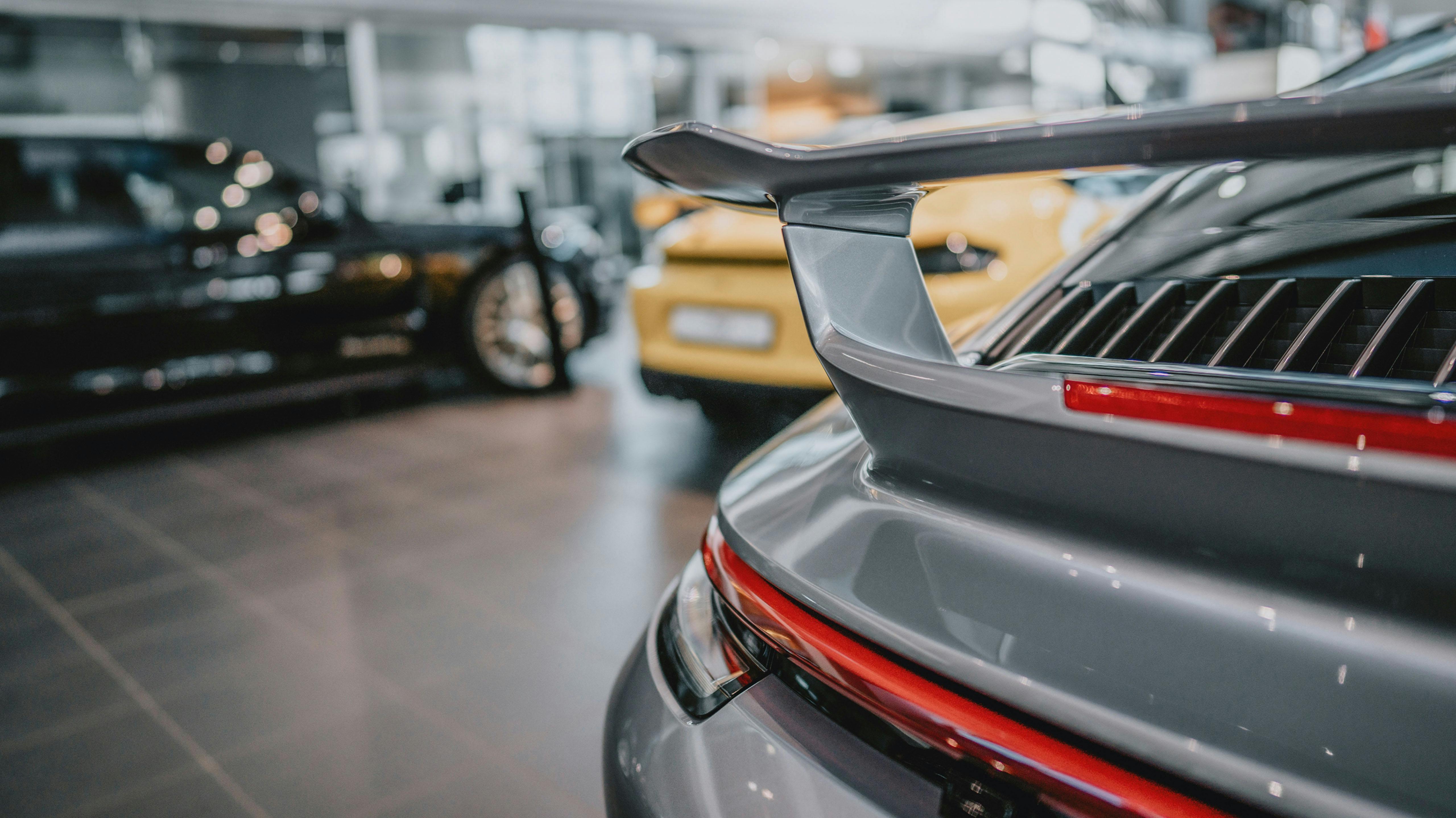 Rapproché du spoiler arrière d'une voiture Porsche gris foncé dans le showroom du Porsche Zenter Lëtzebuerg, avec des voitures Porsche floues visibles en arrière-plan.