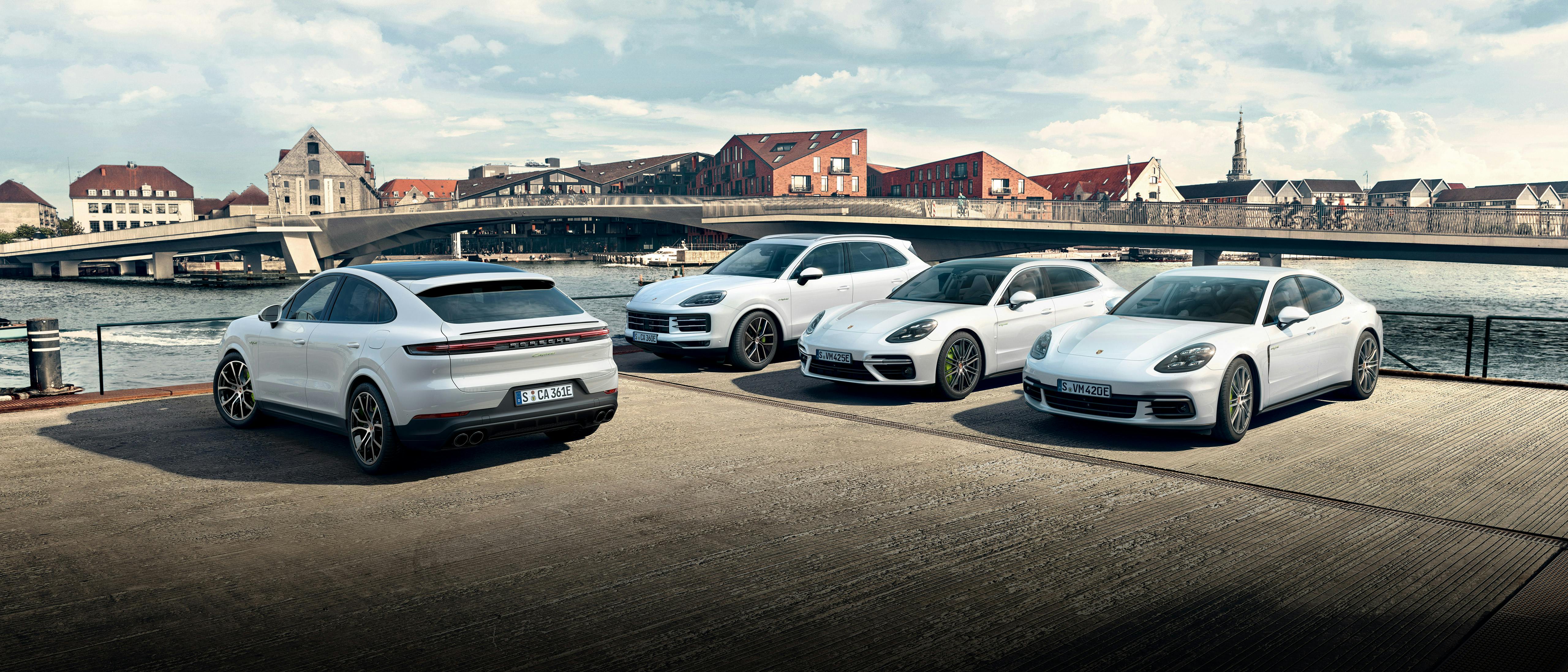Vier weiße Porsche-Modelle (Cayenne und Panamera) stehen nebeneinander vor einer Brücke in einer Stadt mit historischem Flair.