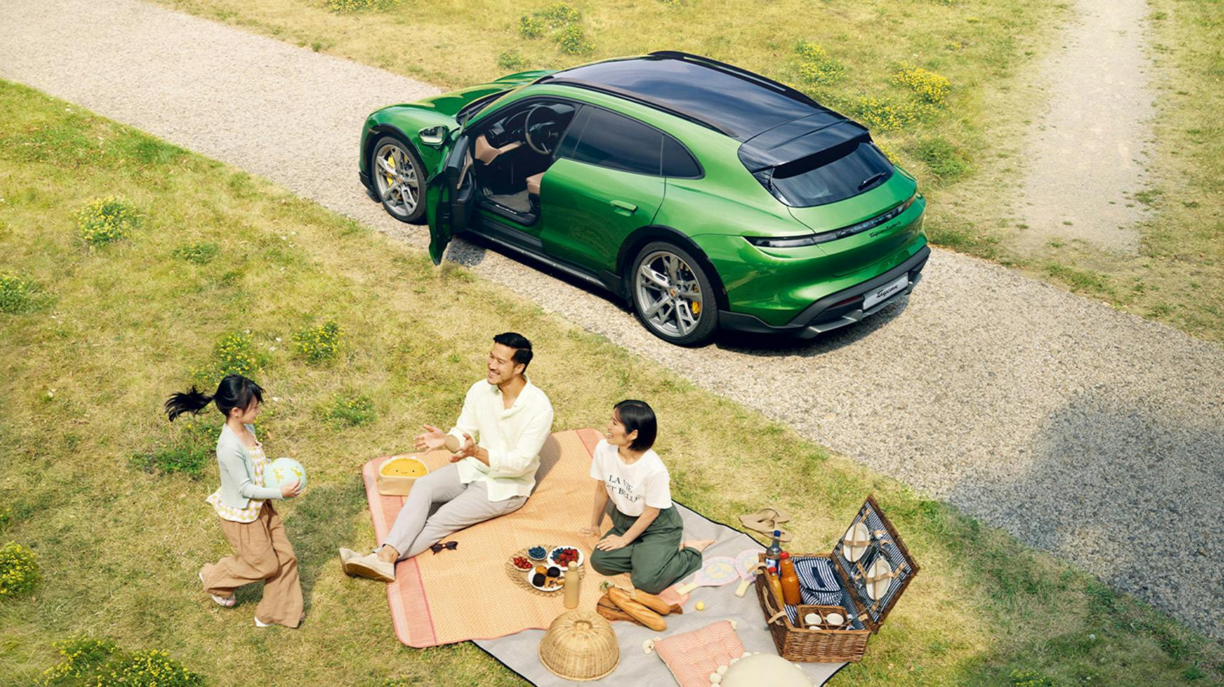 Three people enjoy a picnic on a grassy area near a green Porsche Taycan Cross Turismo parked on a gravel path.