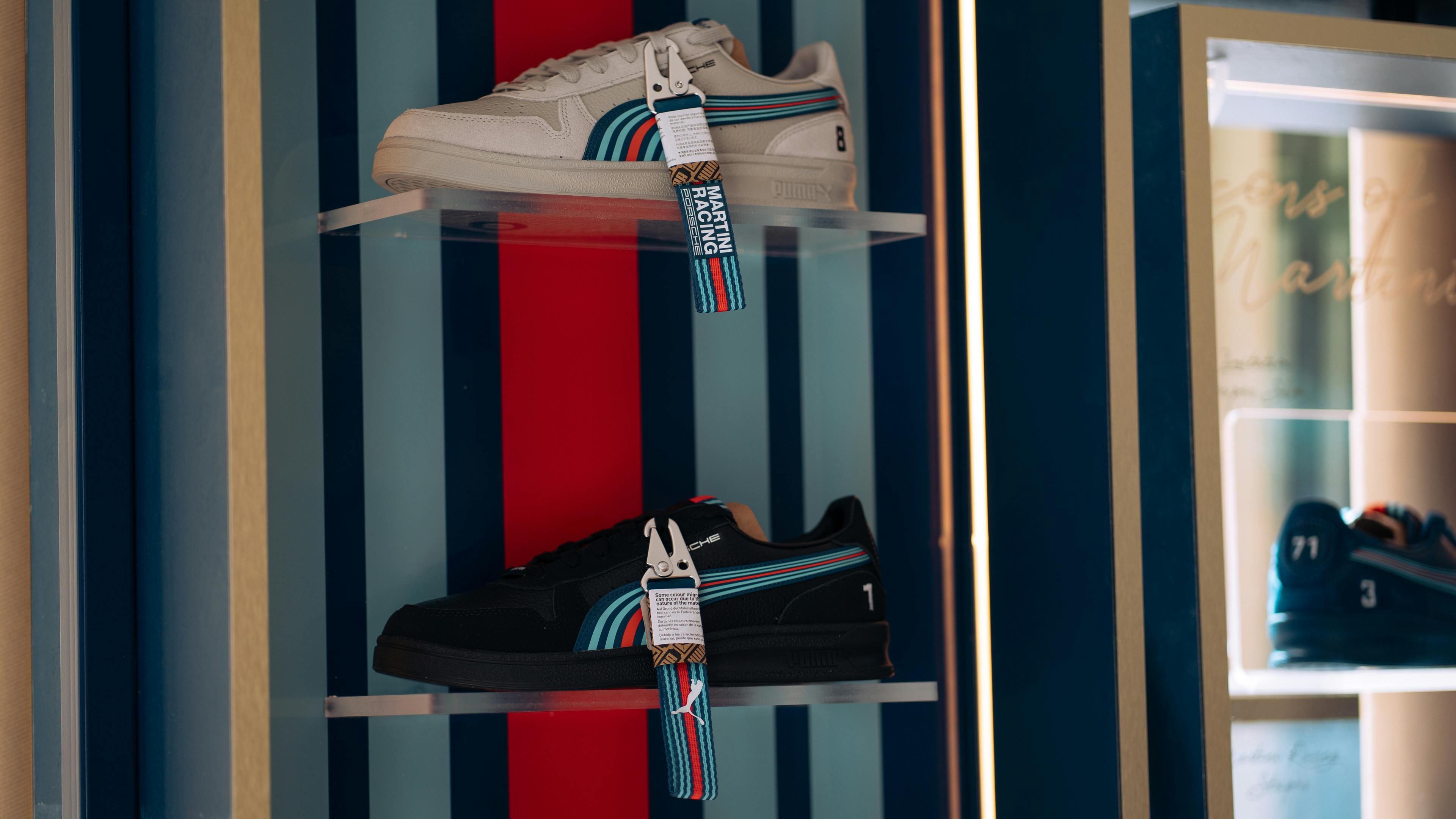 Two Puma x Porsche sneakers from the Martini Racing collection are displayed on clear acrylic shelves against a striped blue and red background. The top sneaker is off-white with teal and red stripes, while the bottom one is black with similar colorful accents. Both shoes feature "Martini Racing" branded tags clipped to the laces.
