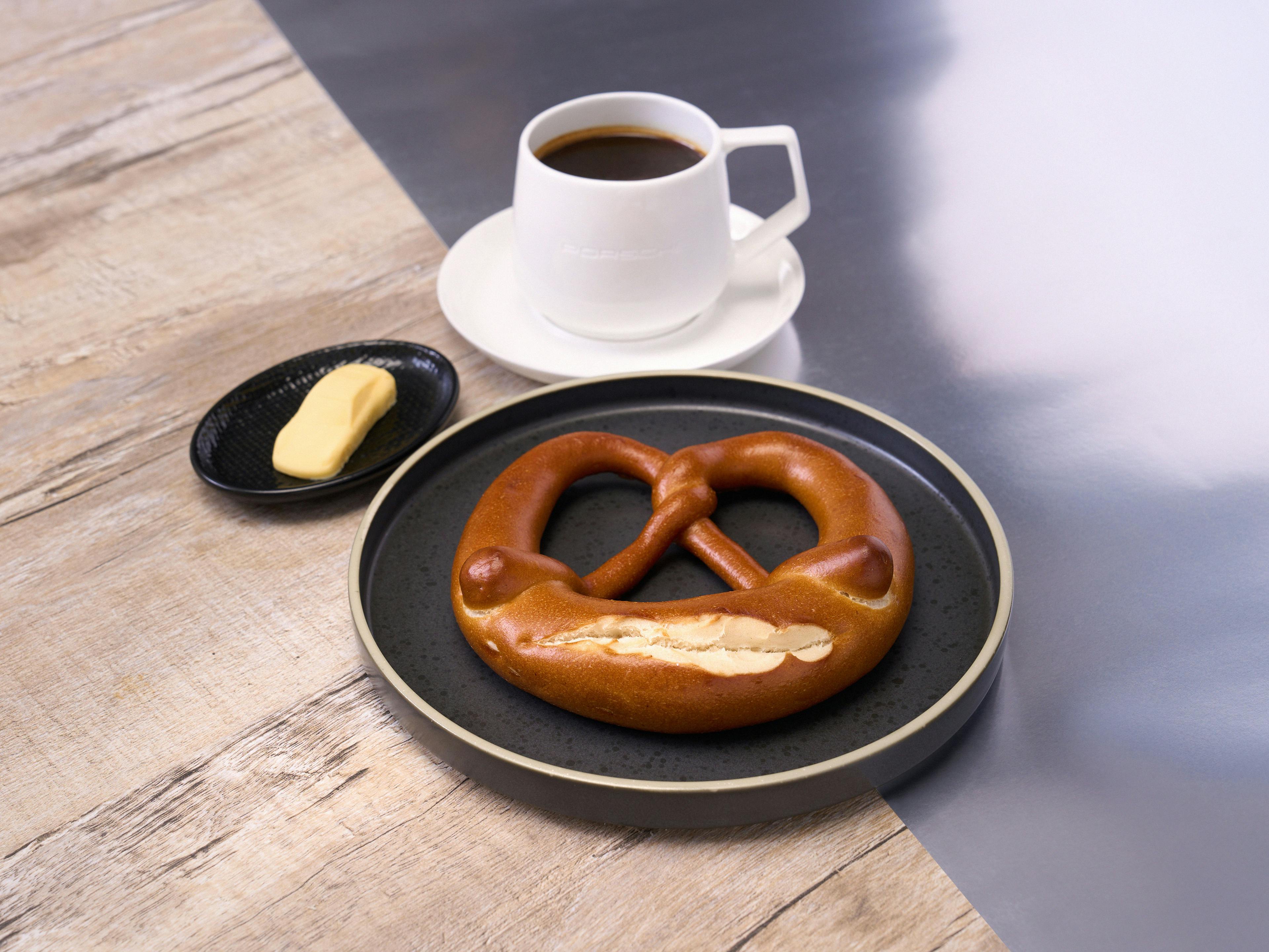 Butter pretzel on black plate with cup of coffee and extra piece of butter on small side plate.