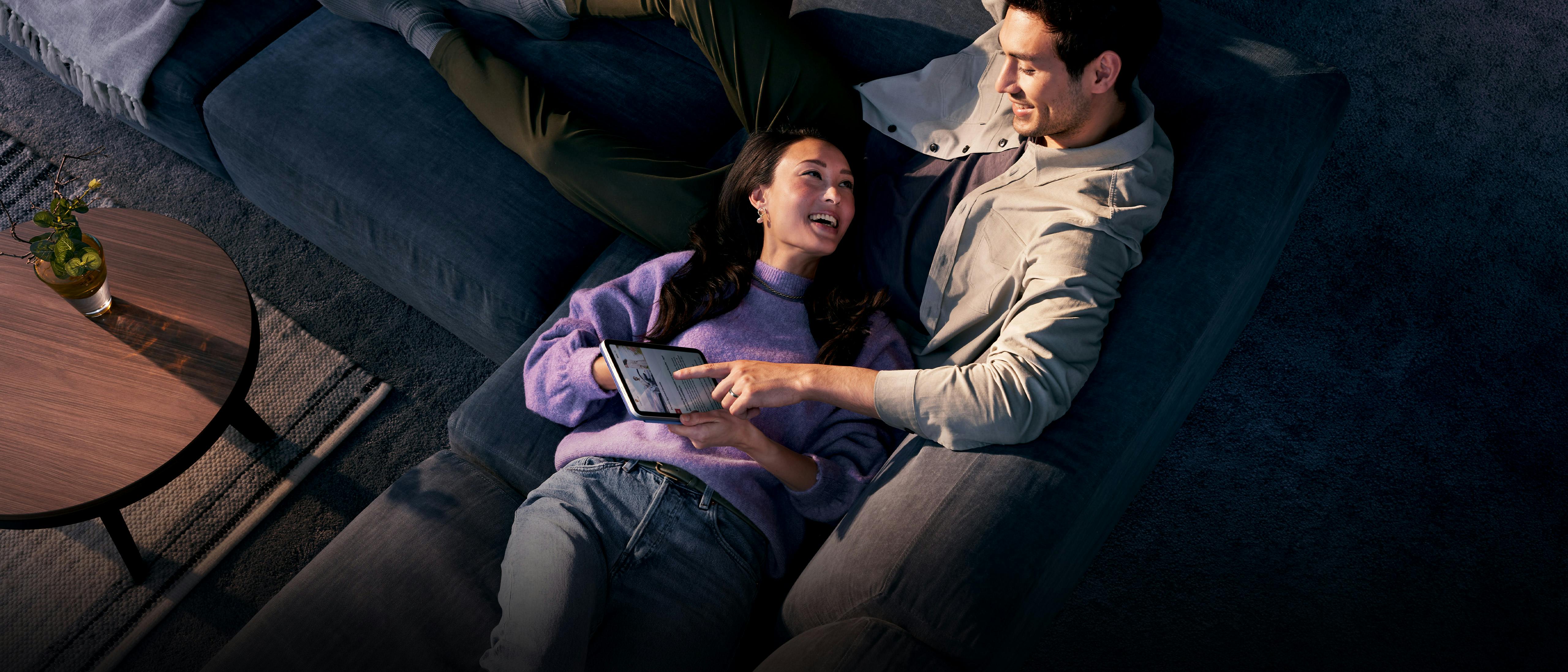 A couple are lying on the sofa. The woman is pointing at the screen of the tablet that the man is holding.