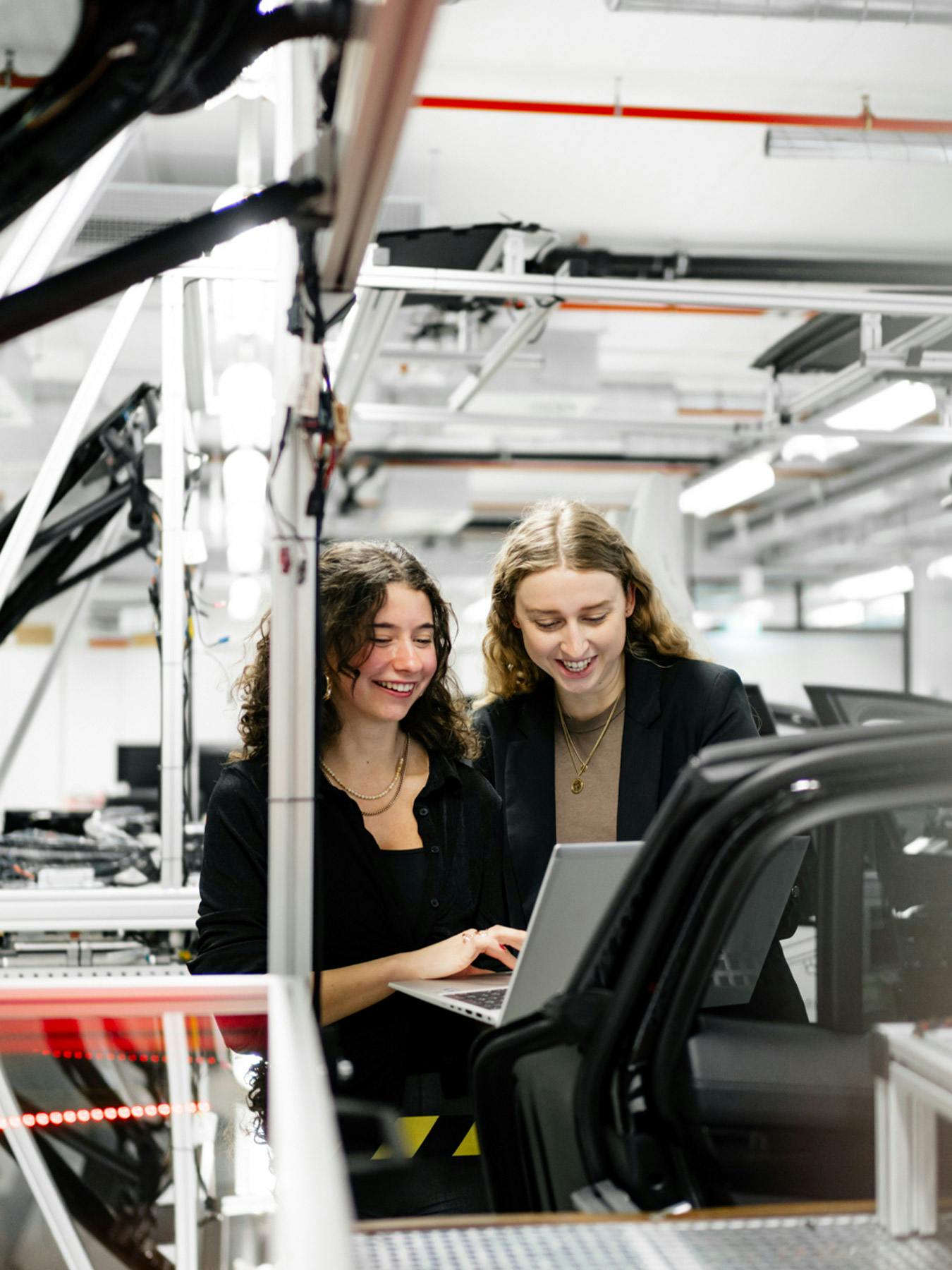 Zwei Studentinnen stehen in einer hellen, technischen Arbeitsumgebung neben einem Fahrzeug und betrachten gemeinsam einen Laptop. Im Vordergrund sind Fahrzeugrahmen und Montageelemente zu sehen, im Hintergrund industrielle Anlagen und Beleuchtung.​