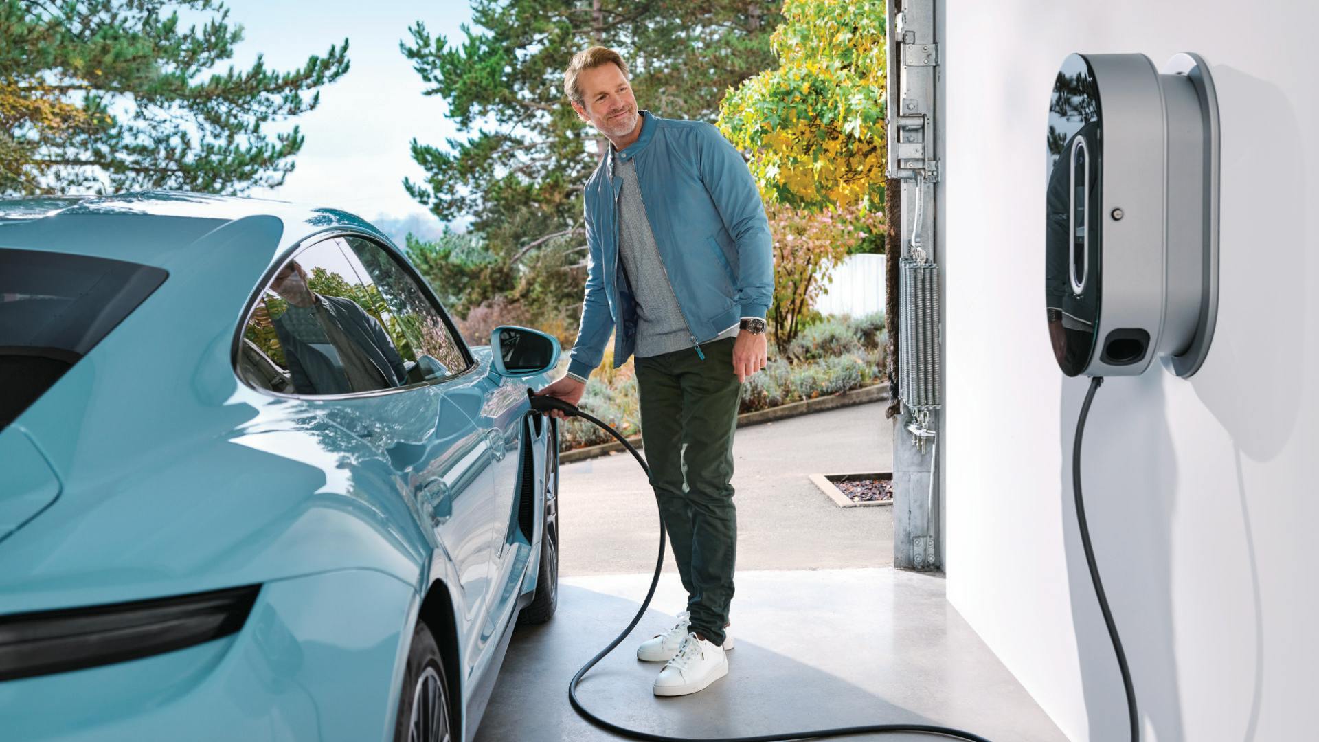 A man in a garage stands next to a Porsche Taycan and charges it via a wall box.