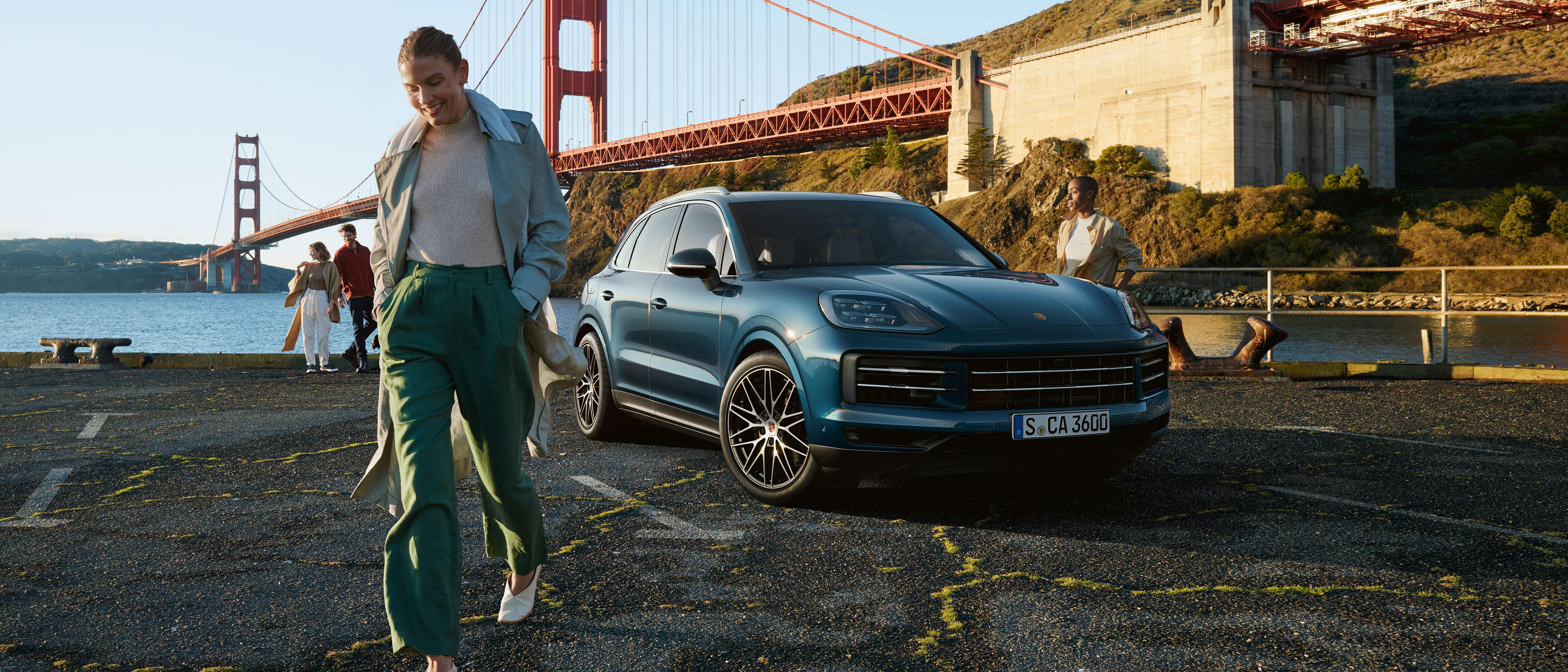 Eine Frau lächelt beim Gehen nahe einem blauen Porsche Cayenne, während die Golden Gate Bridge im Hintergrund zu sehen ist.