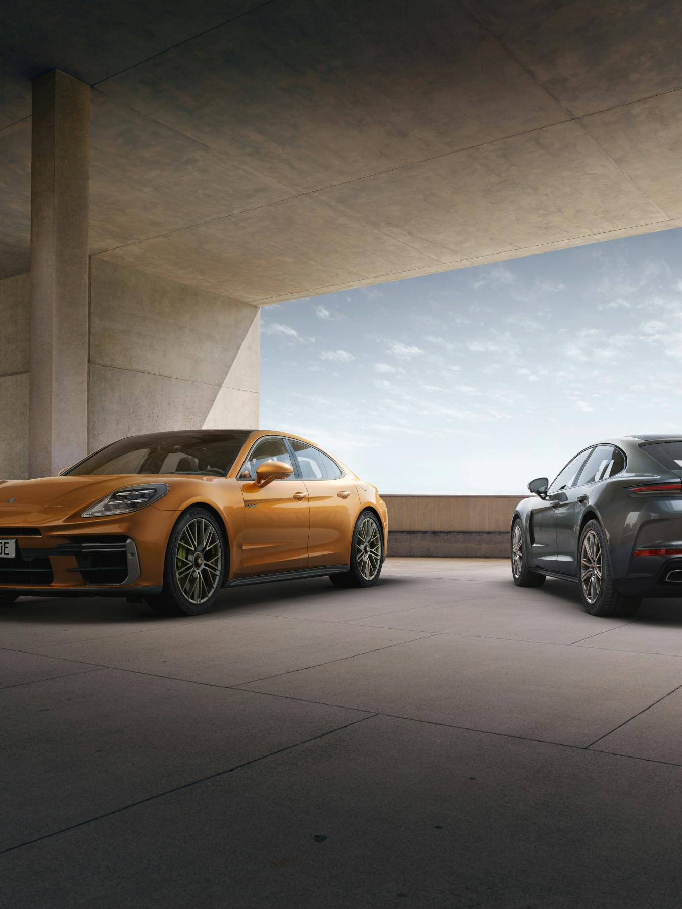 An orange Porsche Panamera Turbo e-Hybrid parked on the left and a grey Porsche Panamera parked on the right on a concrete building. The left car is angled towards the left with the front and the right car towards the right with the back.