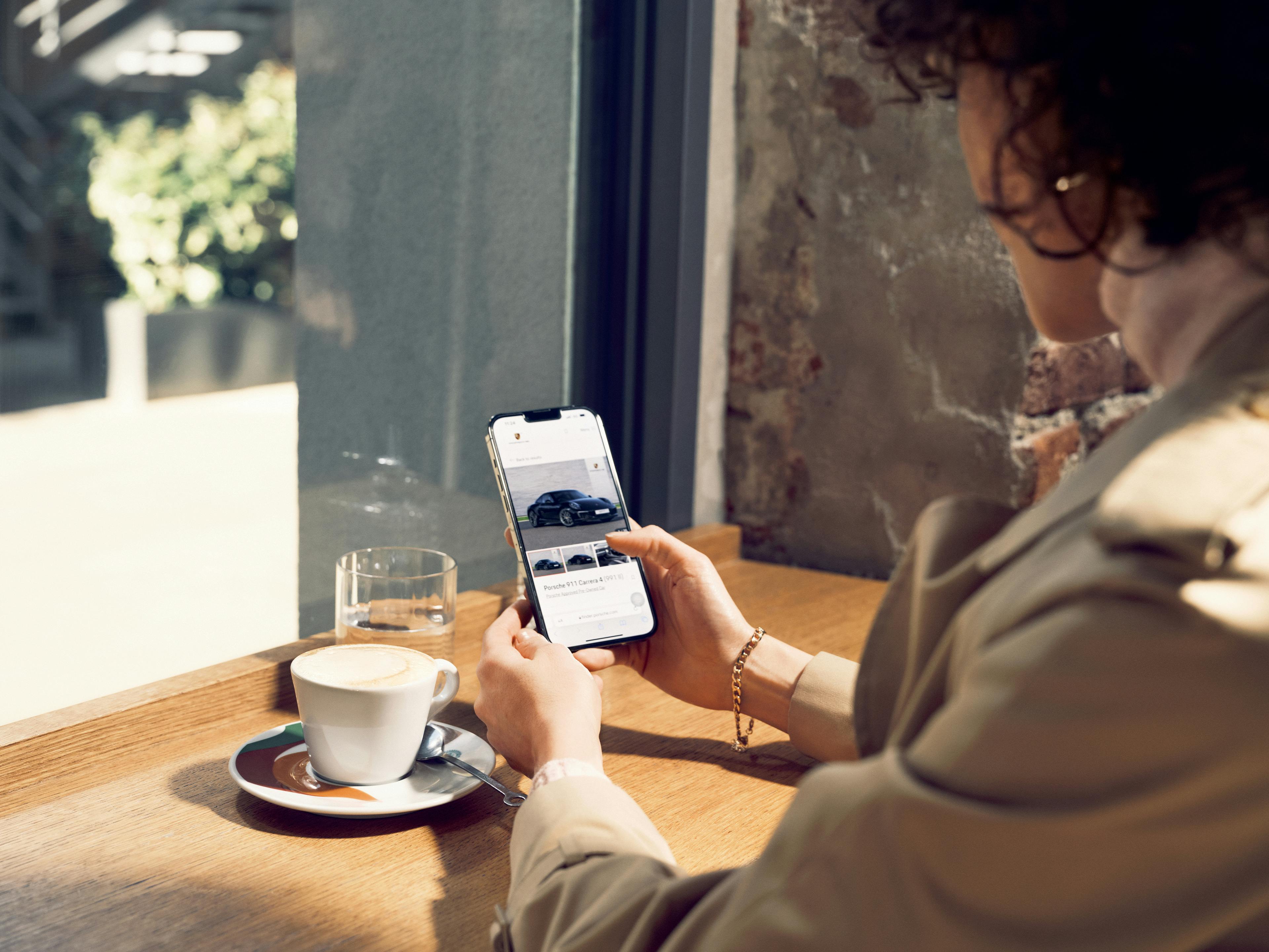 Eine Frau sitzt in einem Café und schaut auf ihr Handy, während die Porsche Approved Pre-owned Car Suche geöffnet ist.