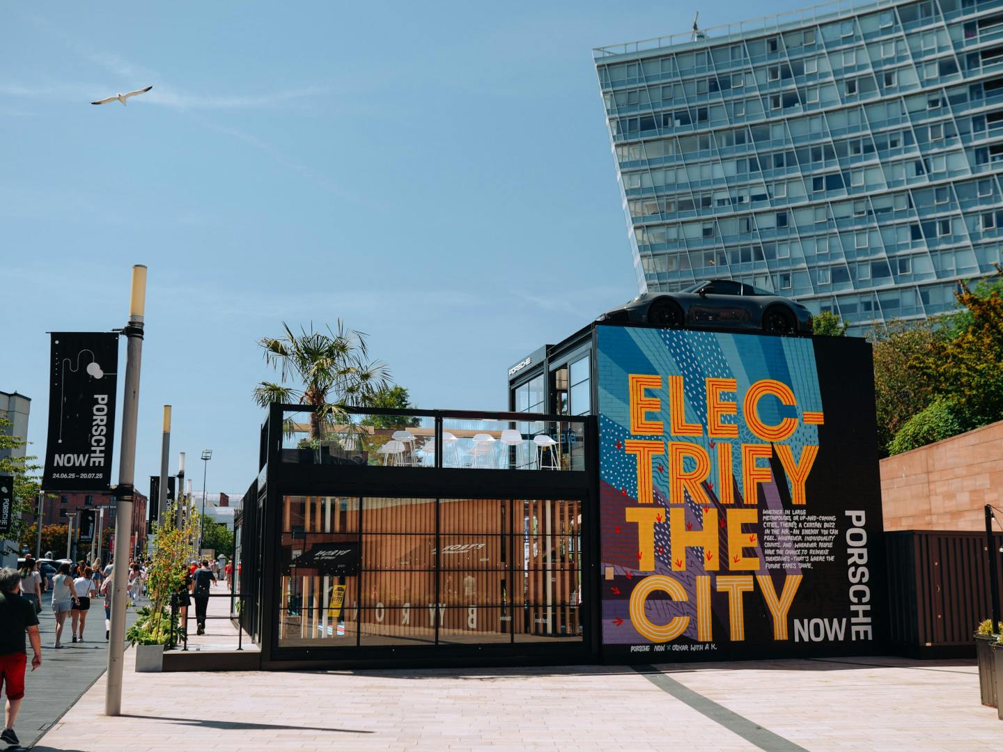 The Porsche Now pop-up in an urban plaza featuring a large sign reading “Electrify the City”, with Porsche sports car displayed on the roof and people walking nearby.