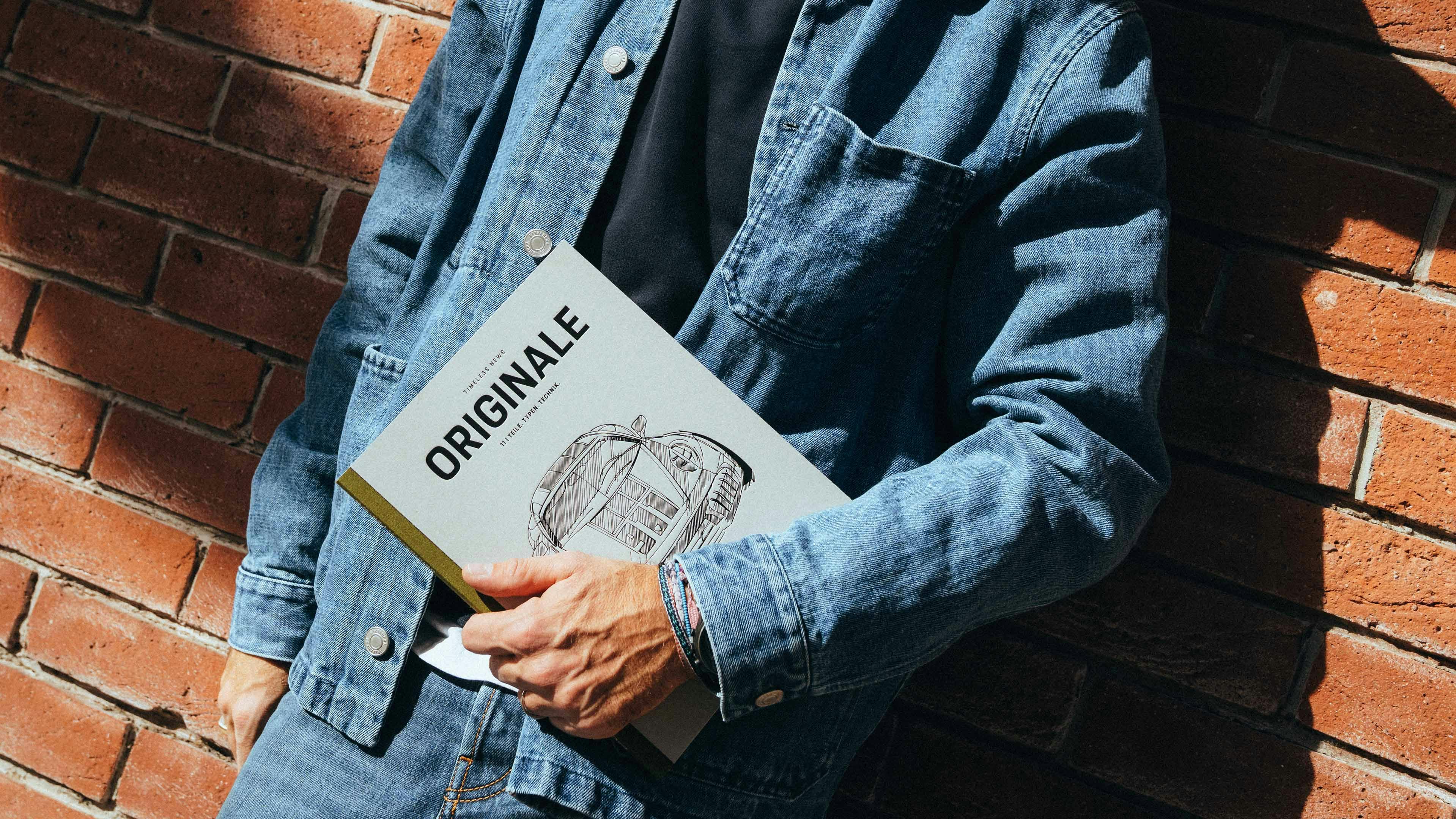 Andrea Vailetti leans against a brick wall holding ORIGINALE 11 in his hand. The cover of the magazine features a drawing of the Spirit 70.