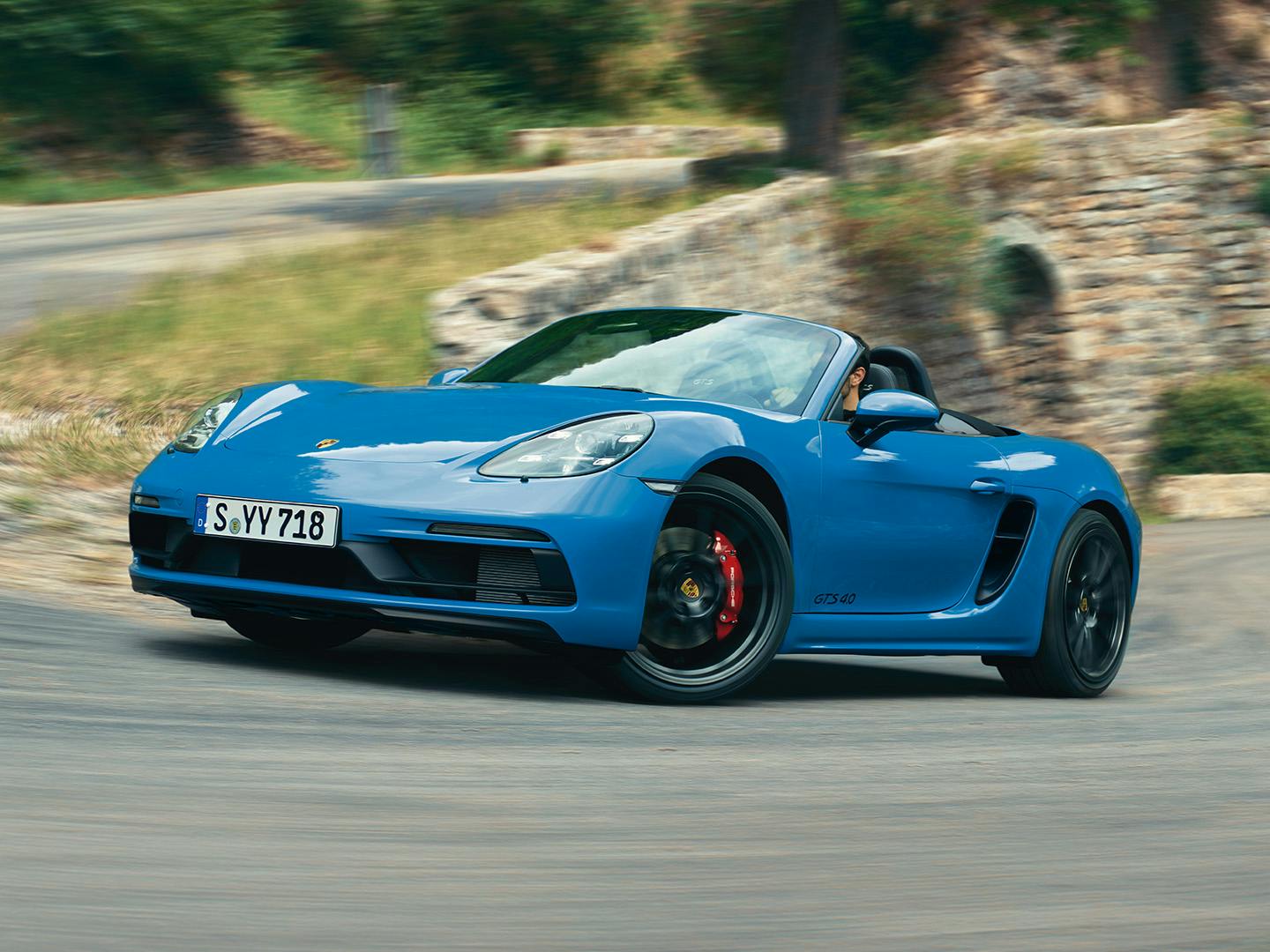 718 Boxster bleue circulant à grande vitesse sur une route de montagne sinueuse bordée d’arbres et de rochers.