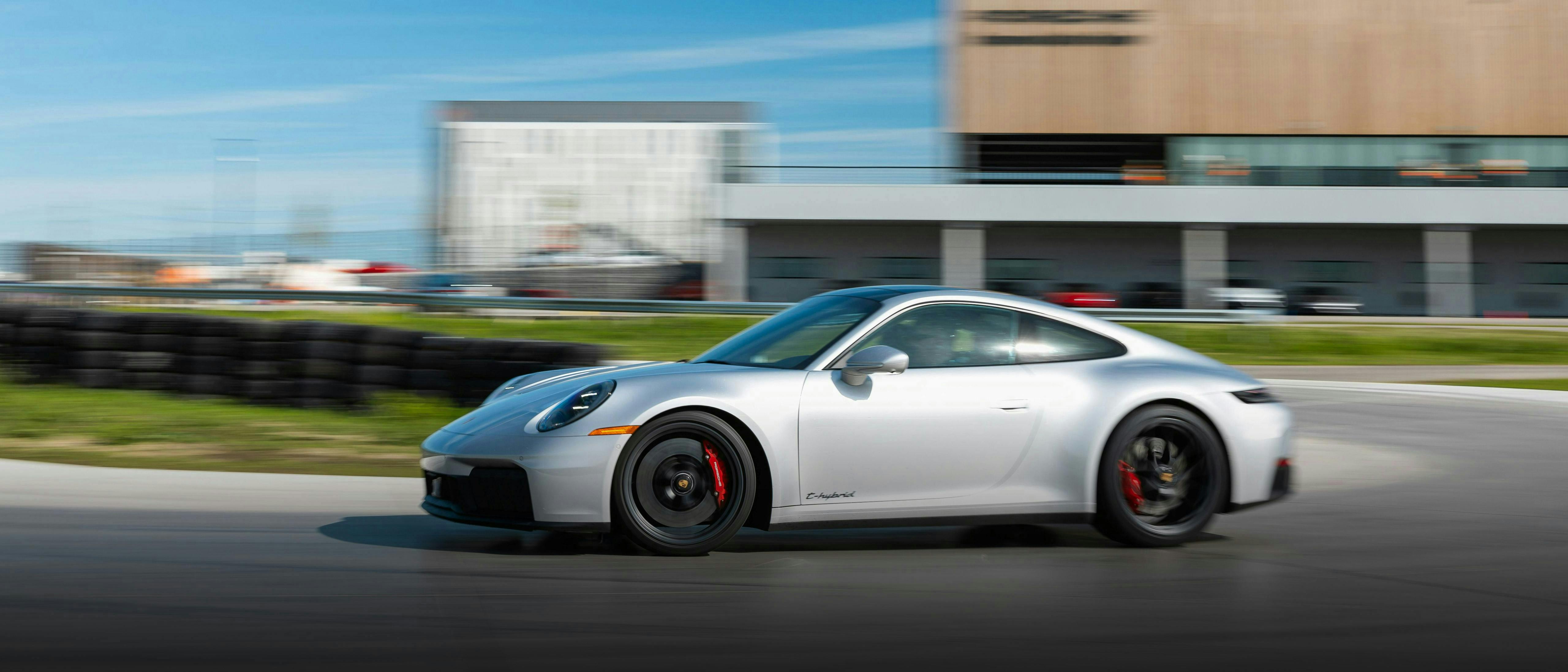 A silver Porsche 911 driving on a polished concrete track with a modern wooden-facade building in the background.