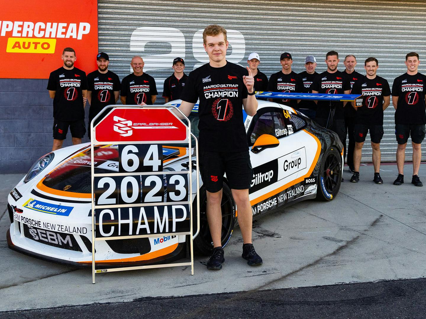 Marco Giltrap stands in front of a Porsche holding his starting and year number as champion, with his team behind him.