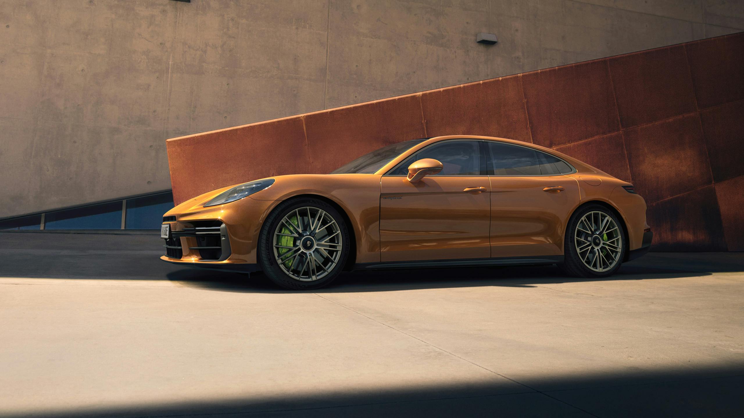 A Panamera Turbo E-Hybrid in Madeira Gold Metallic parked alongside a wall that is copper coloured while there is a lighter brown building behind it.